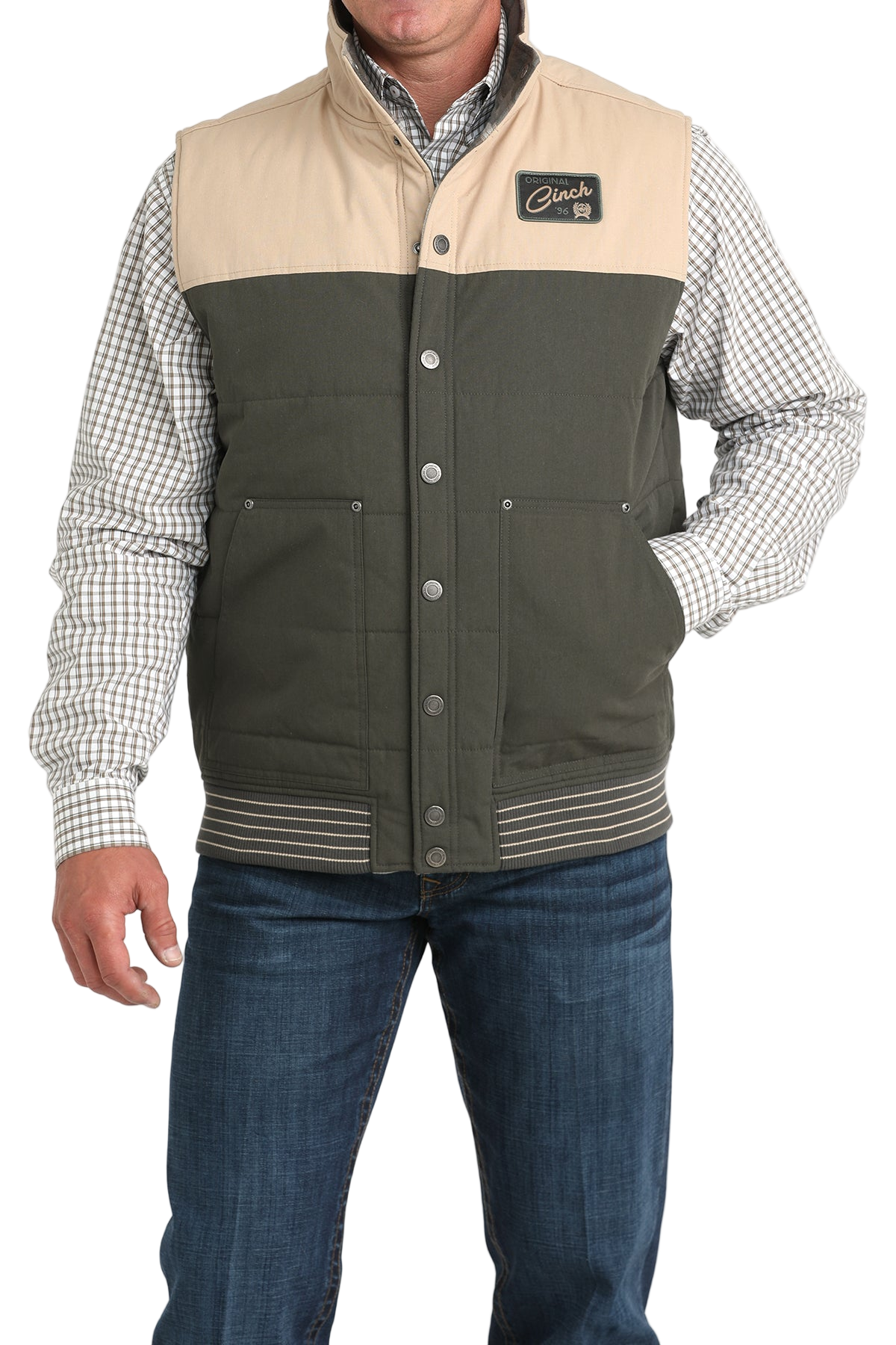 A man wearing a two-tone vest with a beige top panel and dark green lower panel over a checkered button-down shirt, paired with blue jeans