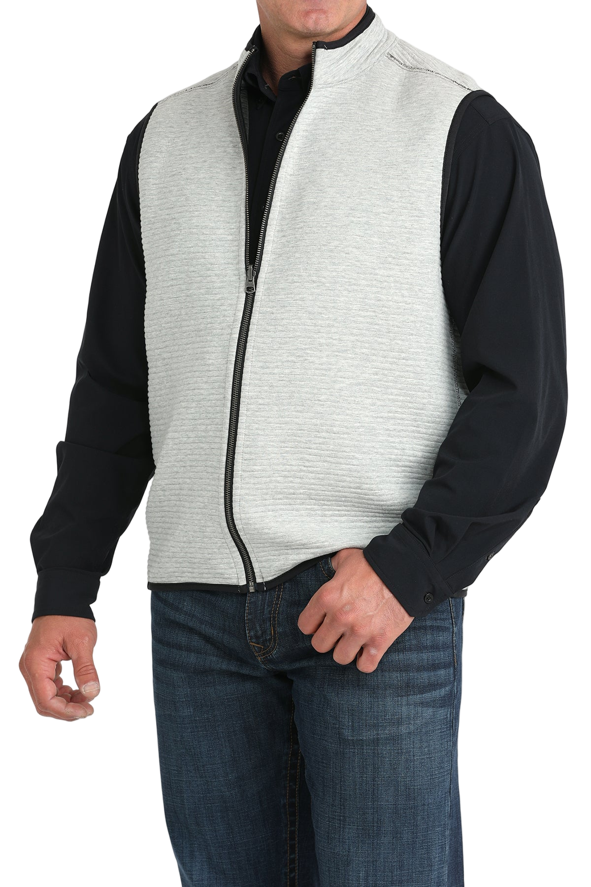 A man wears a light gray zip-front vest over a black long-sleeve shirt and blue jeans