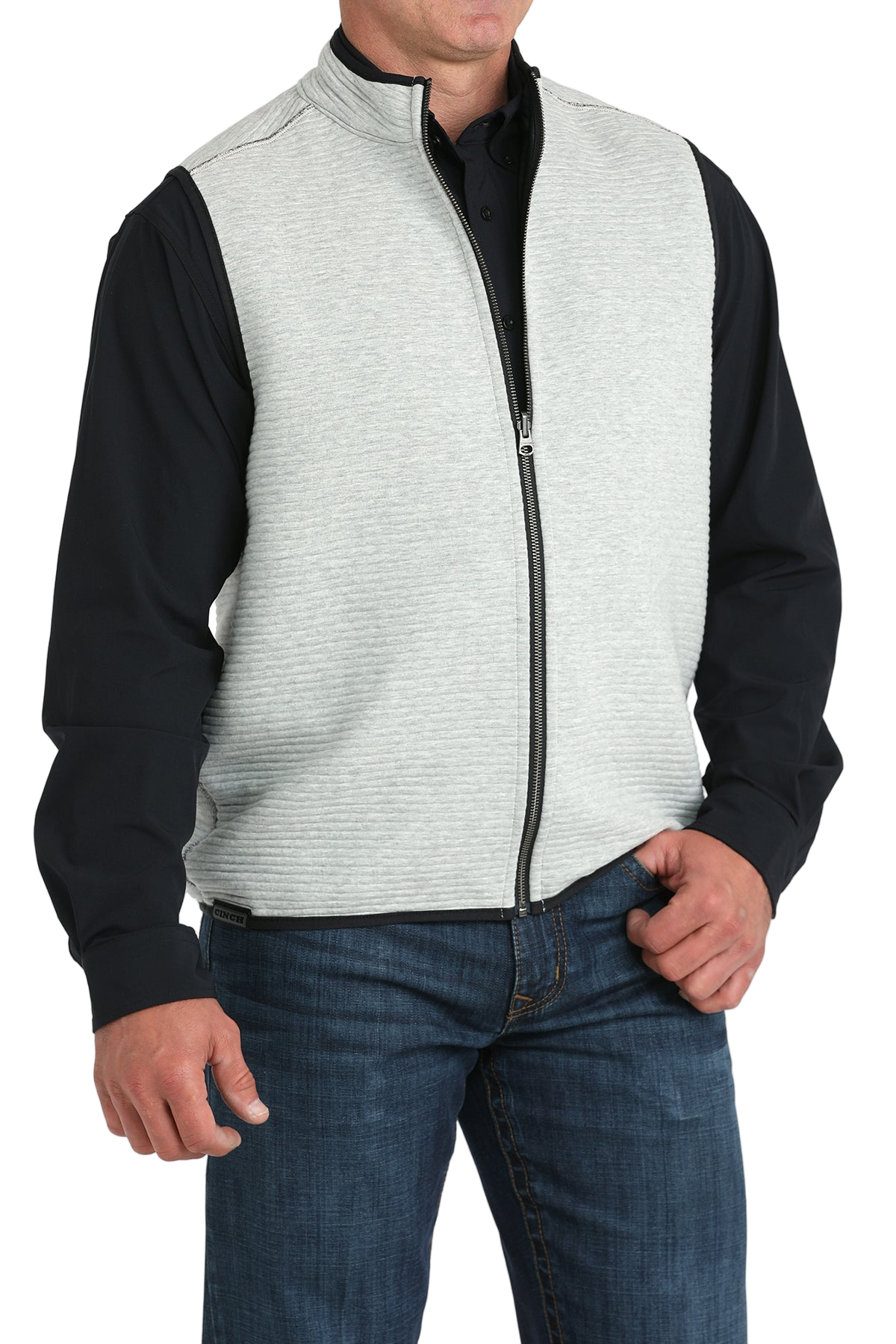 A man wears a light gray zip-front vest over a black long-sleeve shirt and blue jeans