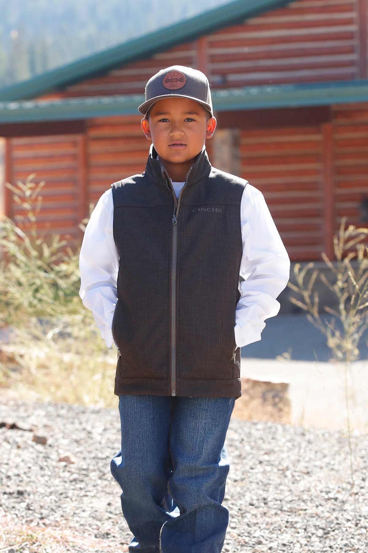 A young boy wearing a gray cap, dark vest, white long-sleeve shirt, and jeans stands outdoors in front of a wooden building