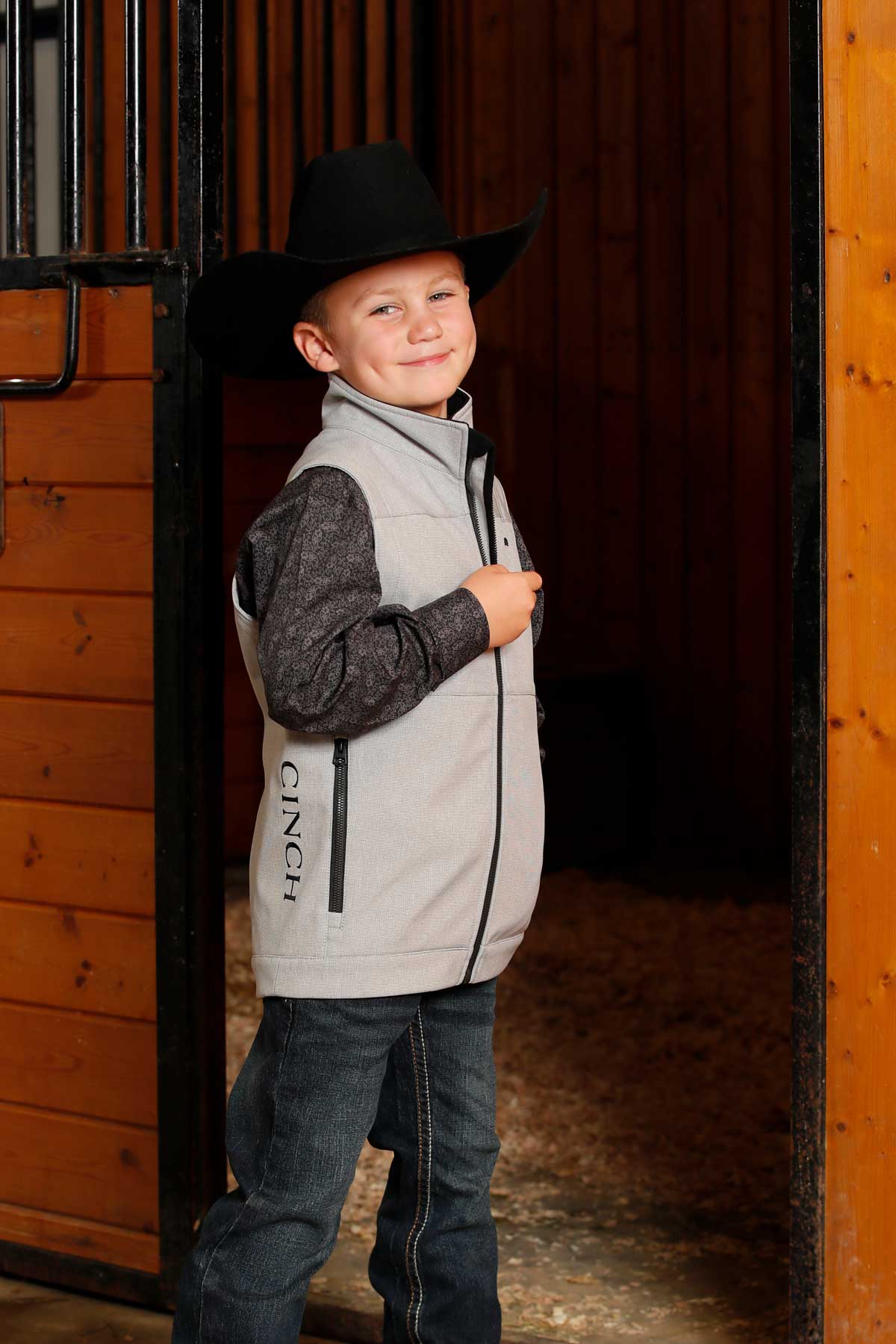 A young boy wearing a black cowboy hat, gray vest, and jeans stands smiling in a barn doorway
