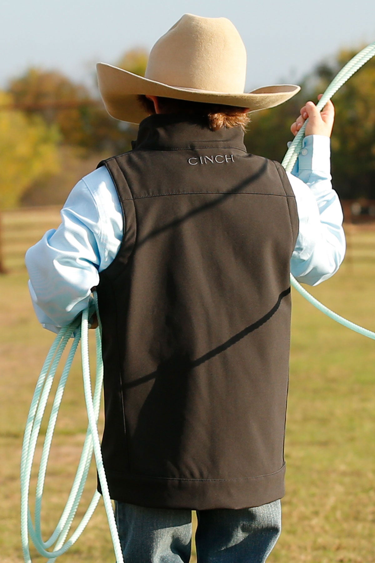 A young boy wearing a cowboy hat, light blue shirt, and black vest stands outdoors holding a rope, shown from the back