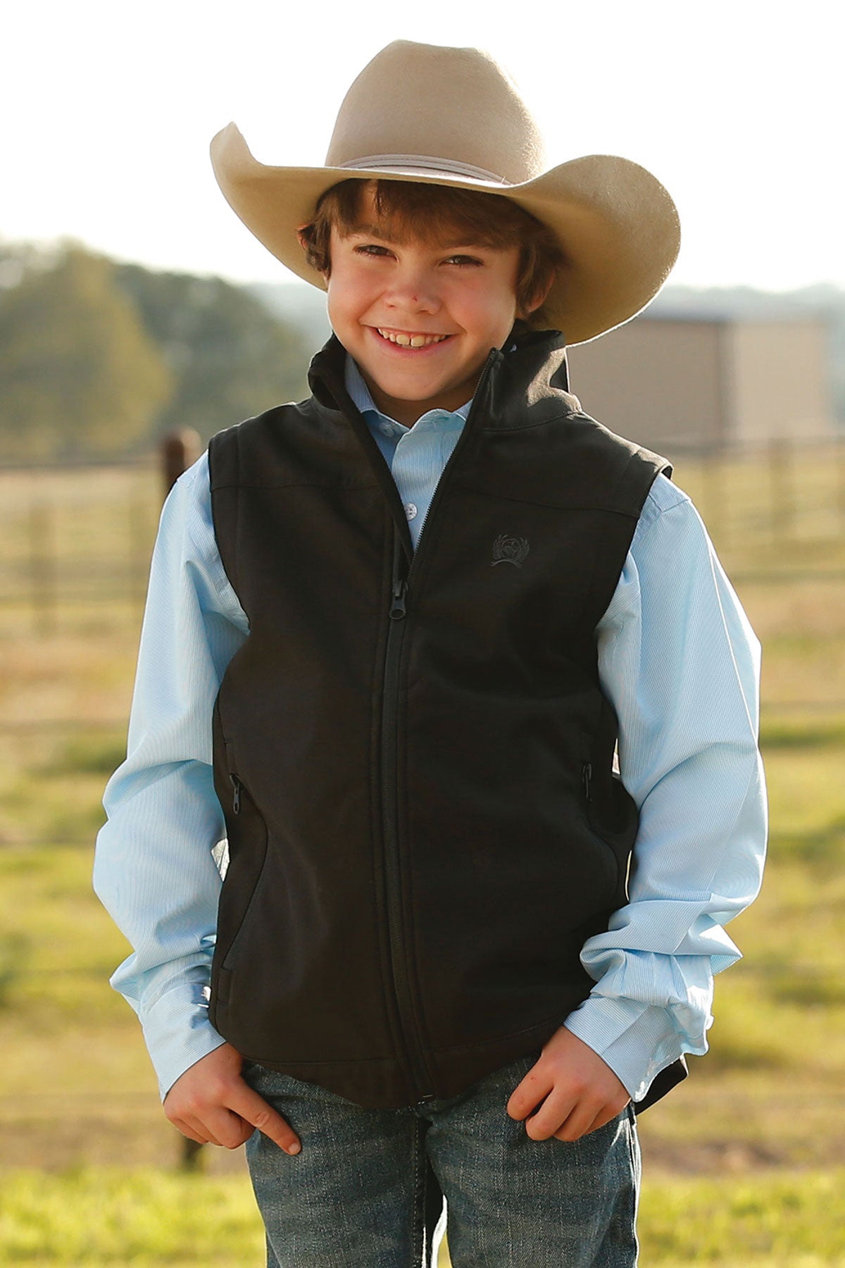 A young boy wearing a cowboy hat, light blue shirt, and black vest stands outdoors smiling