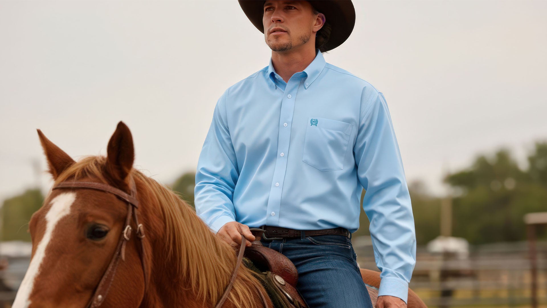 Man riding a horse wearing a light blue long sleeve button-down, jeans and a brown cowboy hat