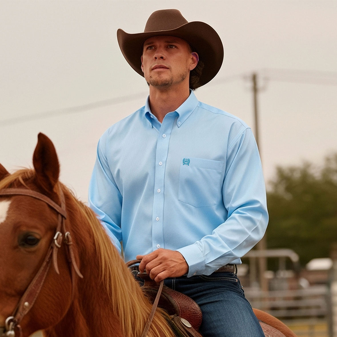 Man riding a horse wearing a light blue long sleeve button-down, jeans and a brown cowboy hat