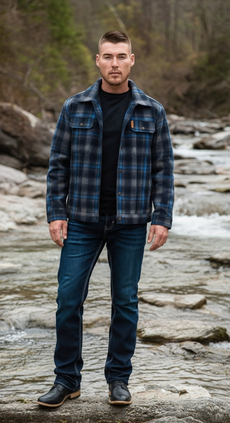 Man standing on rocks near a flowing river, wearing a blue plaid jacket, black shirt, dark wash jeans, and black boots, with trees in the background