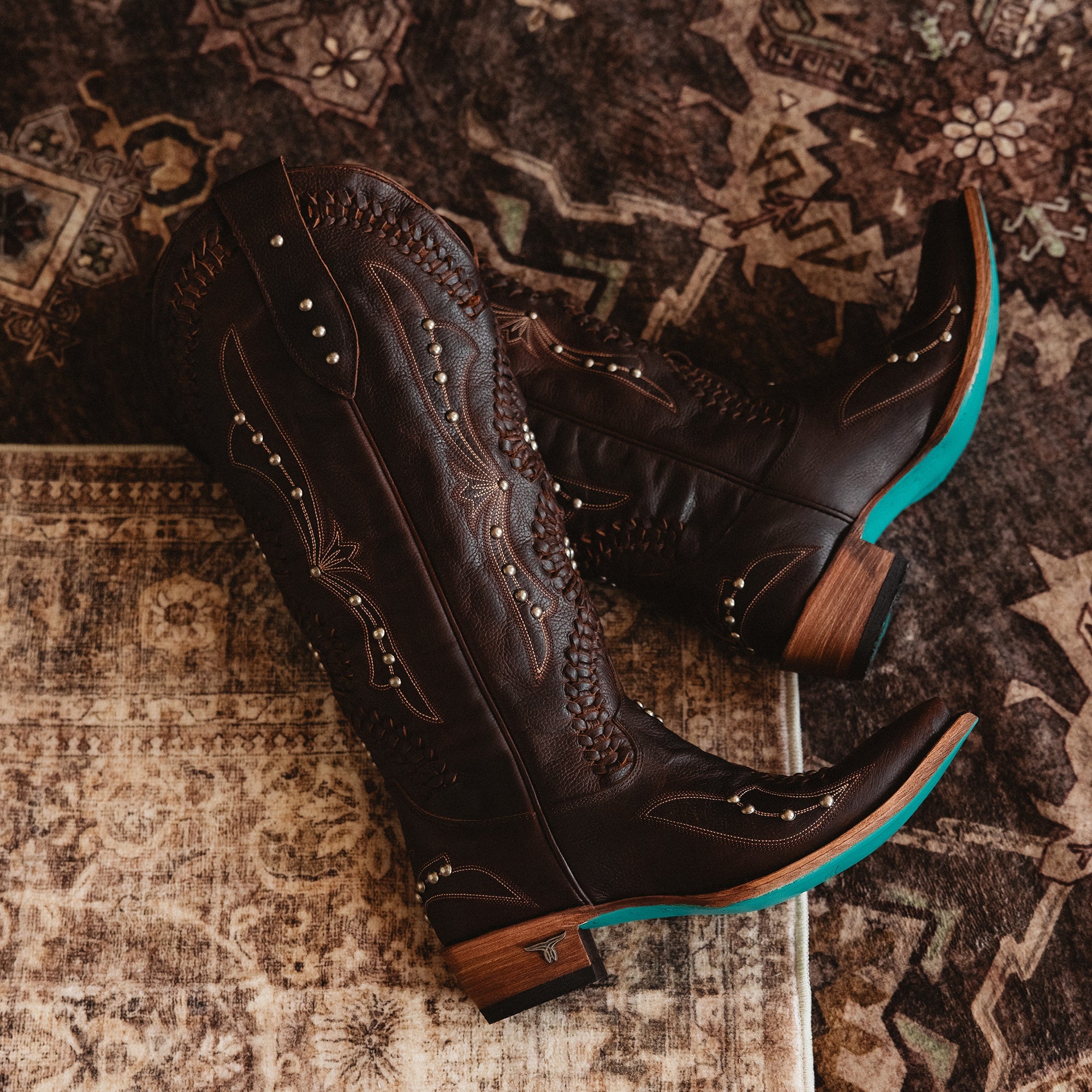 Pair of dark brown cowboy boots with intricate stitching, silver stud details, wooden heels, and turquoise soles, placed on a patterned rug