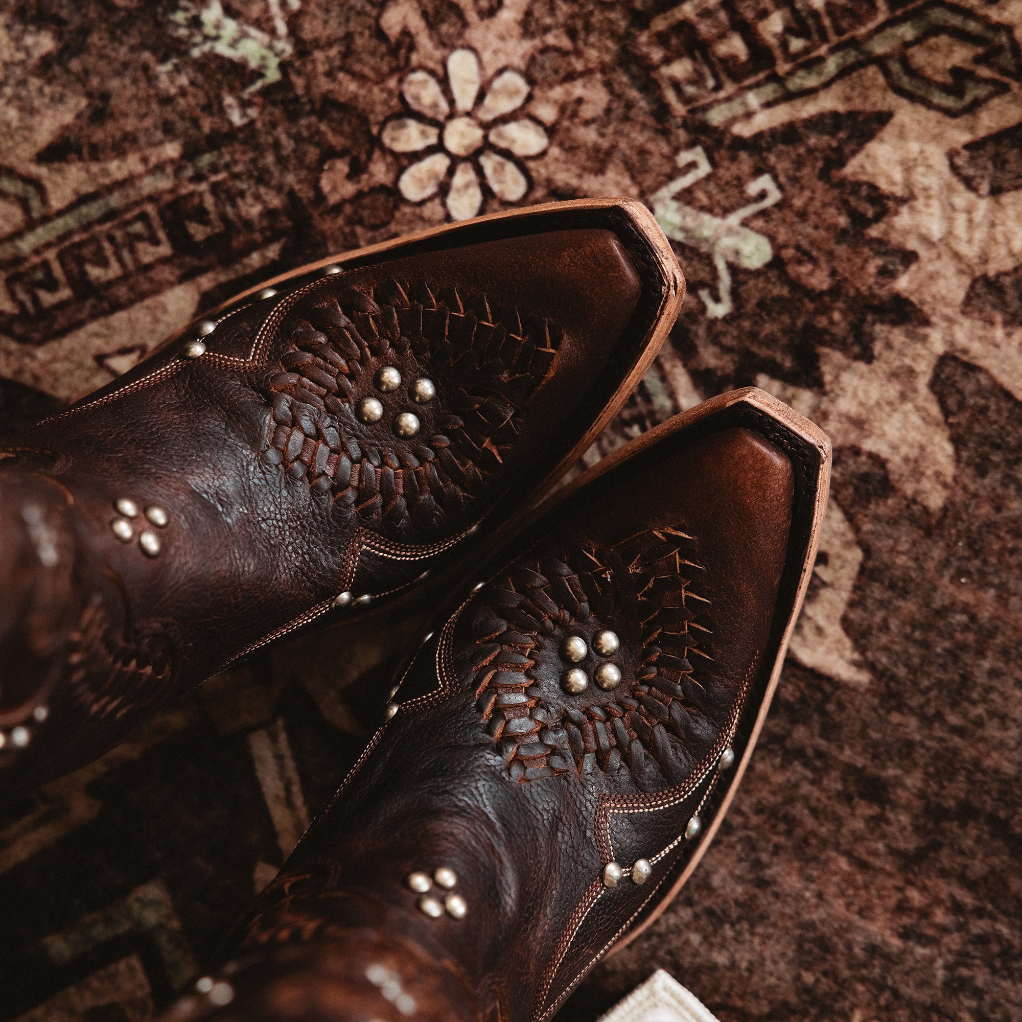 Close-up top view of dark brown cowboy boots with detailed stitching, silver stud accents, and snip toes on a patterned rug