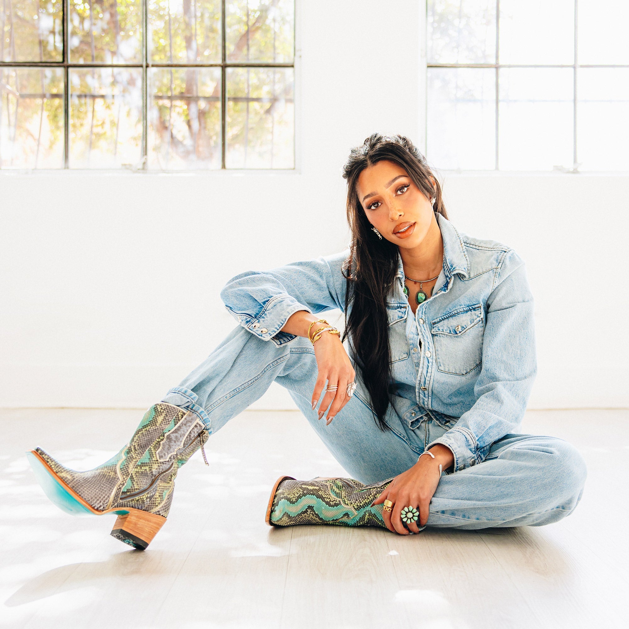 Woman sitting on a bright floor with legs extended and crossed, wearing a light denim jumpsuit and short snakeskin cowboy boots in shades of gray, brown, and turquoise. The boots feature a wooden heel and turquoise sole