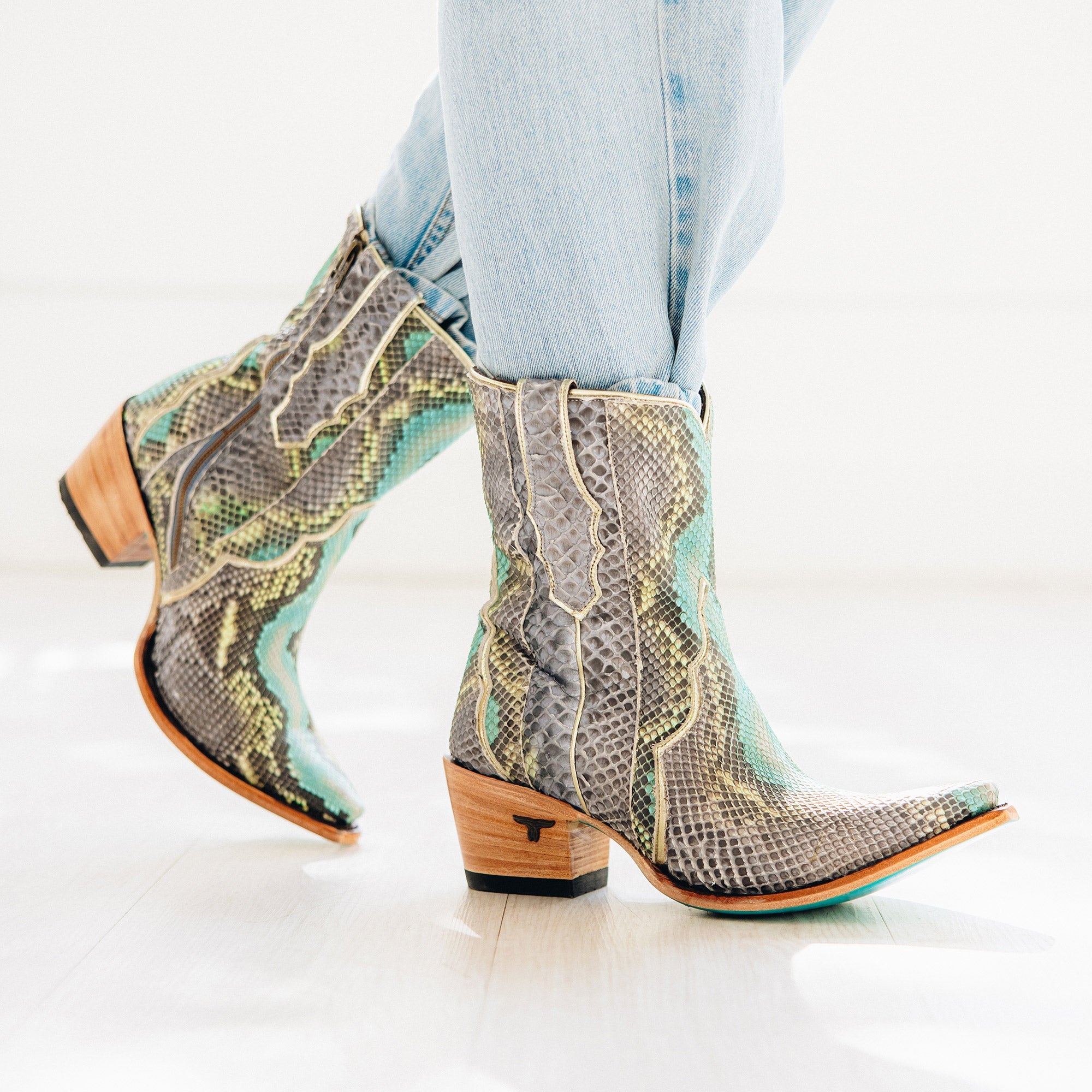 Pair of short cowboy boots with a bold snakeskin pattern in shades of gray, turquoise, and cream. The boots feature a snip toe, stacked wooden heel, and turquoise soles for a striking contrast