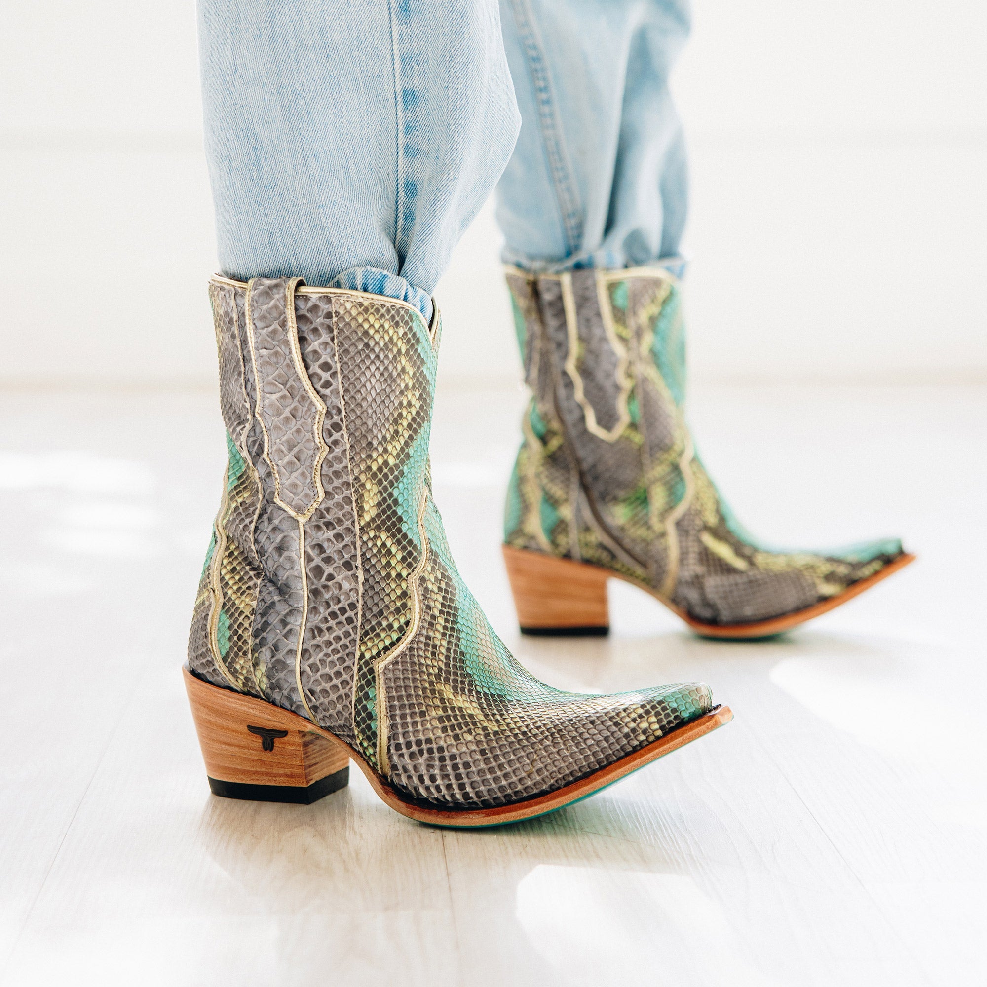 Pair of short cowboy boots with a bold snakeskin pattern in shades of gray, turquoise, and cream. The boots feature a snip toe, stacked wooden heel, and turquoise soles for a striking contrast