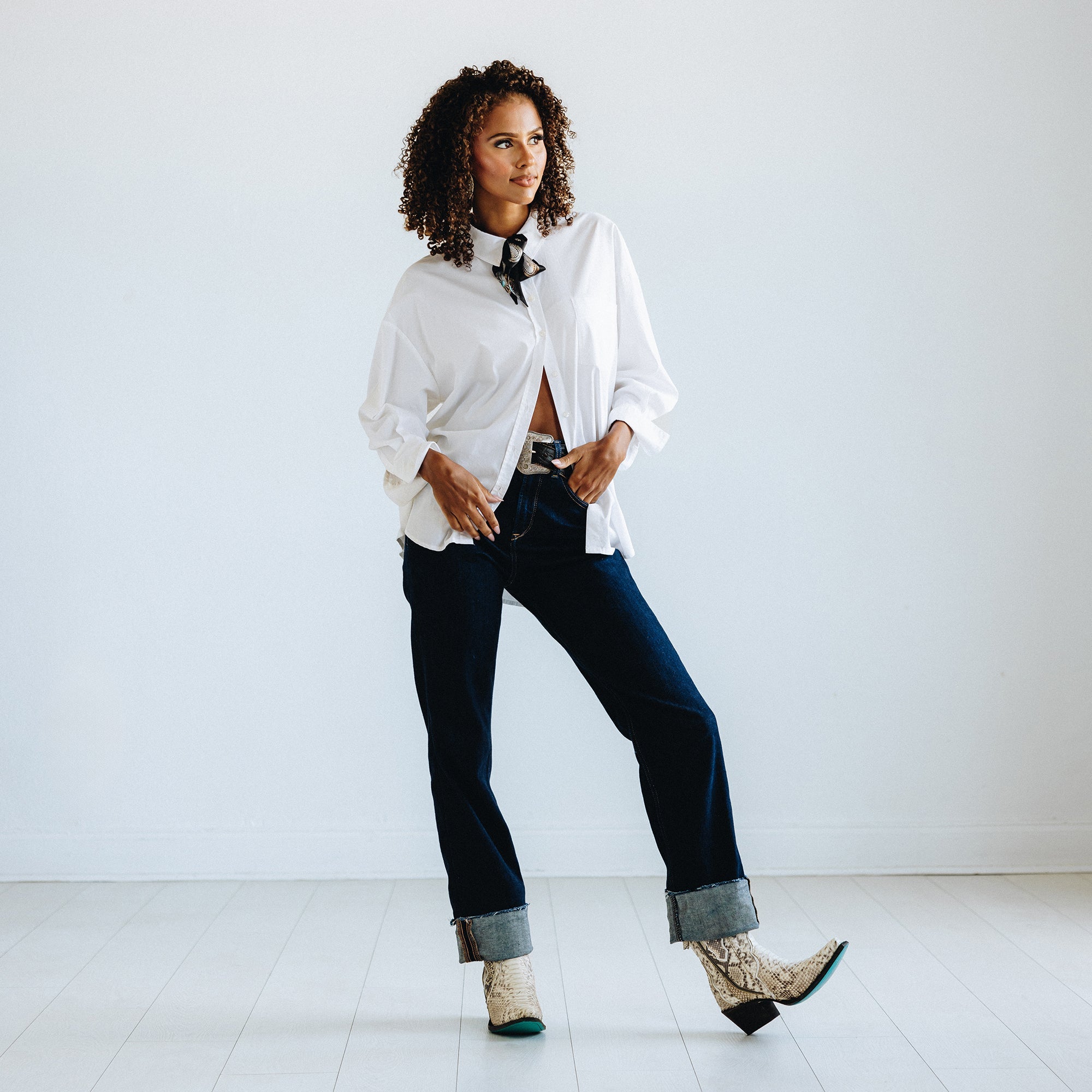Woman standing in a minimal white room wearing a loose white button-down shirt, dark cuffed jeans, and turquoise snakeskin cowboy boots