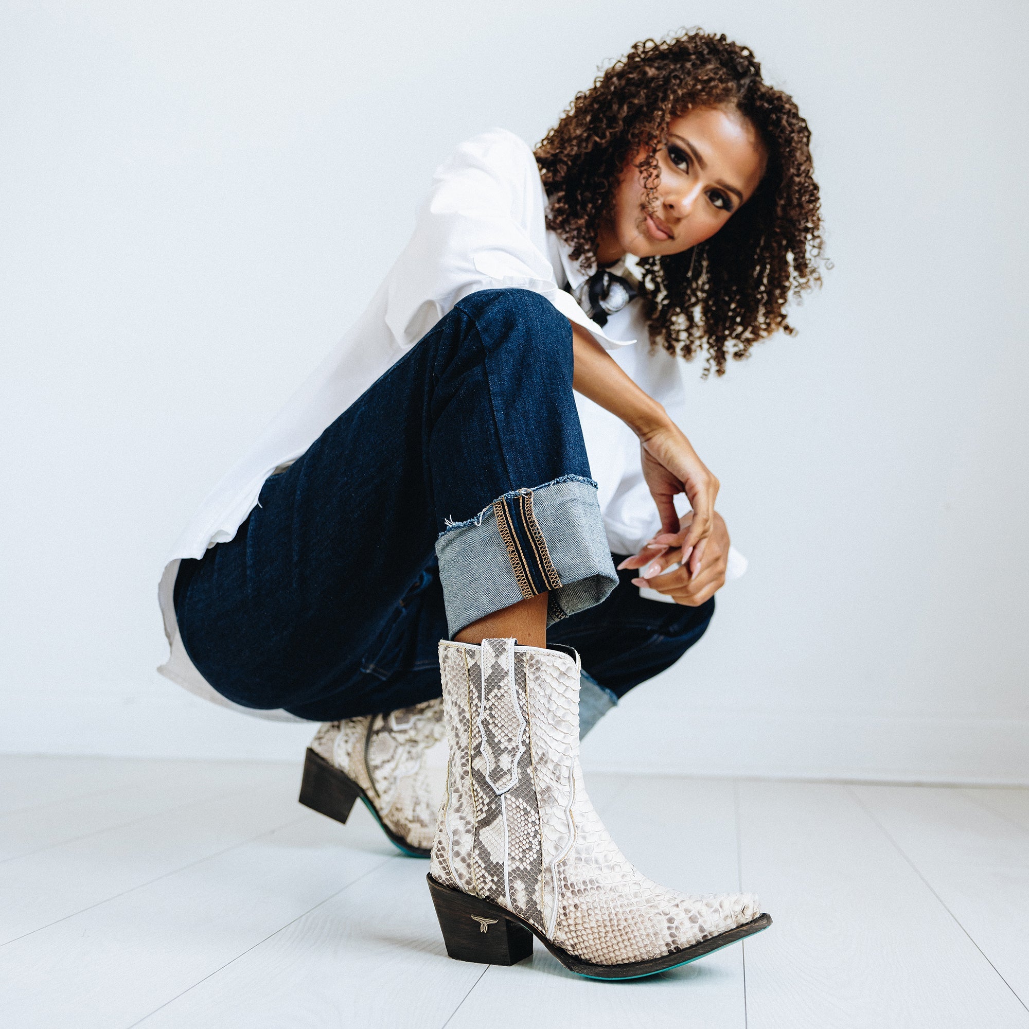 Woman crouching in a minimal white room wearing a loose white button-down shirt, dark cuffed jeans, and turquoise snakeskin cowboy boots