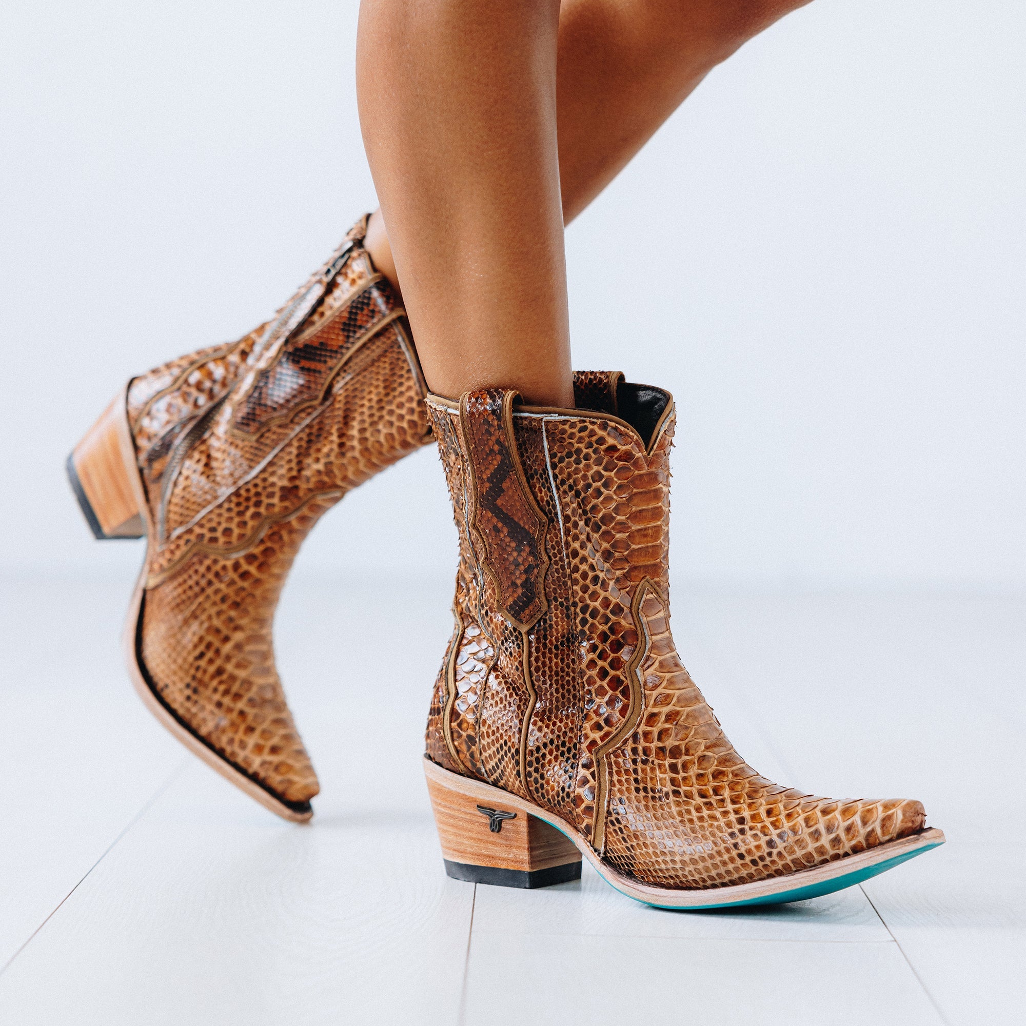 Close-up of a person wearing brown snakeskin cowboy boots with turquoise soles and wooden heels. The boots feature a snip toe and stitched detailing