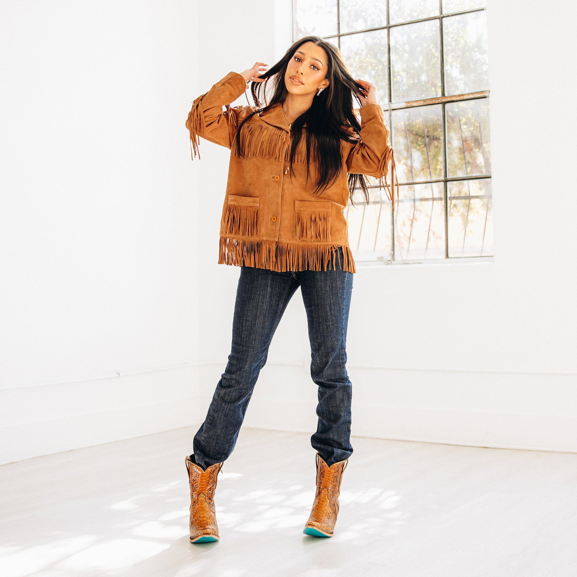 Woman standing in a bright room wearing a tan fringe jacket, dark denim jeans, and brown snakeskin cowboy boots with turquoise soles. She has her hands lifted to her hair and looks confidently toward the camera