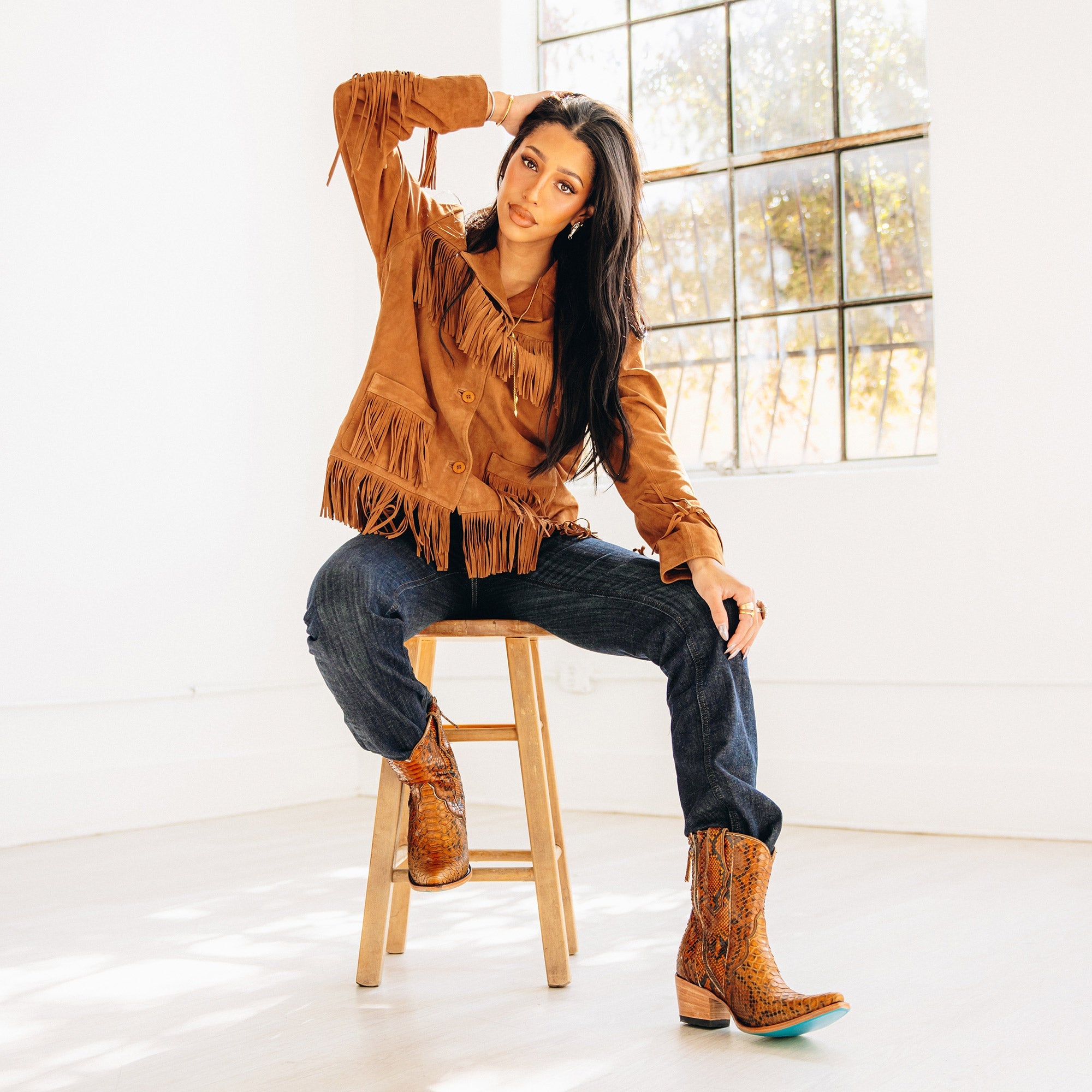 Woman sitting on a wooden stool in a bright room, wearing a brown fringe jacket, dark denim jeans, and brown snakeskin cowboy boots with turquoise soles. She has one hand resting on her head and looks toward the camera