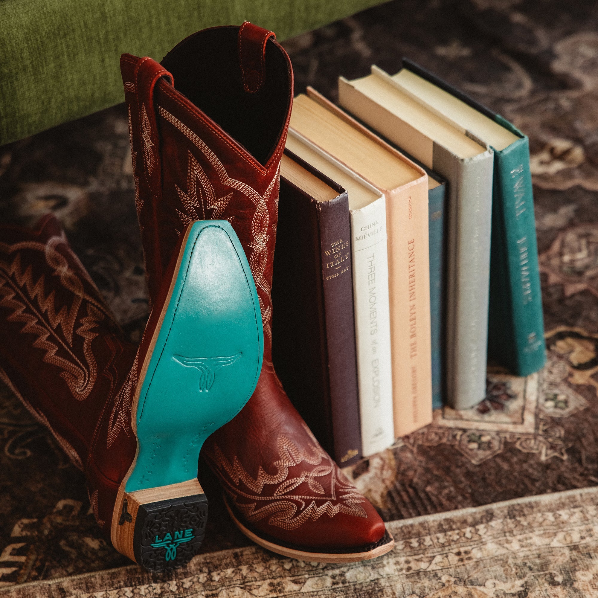 Pair of red cowboy boots with decorative stitching and turquoise soles, placed on a patterned rug leaning against a stack of books