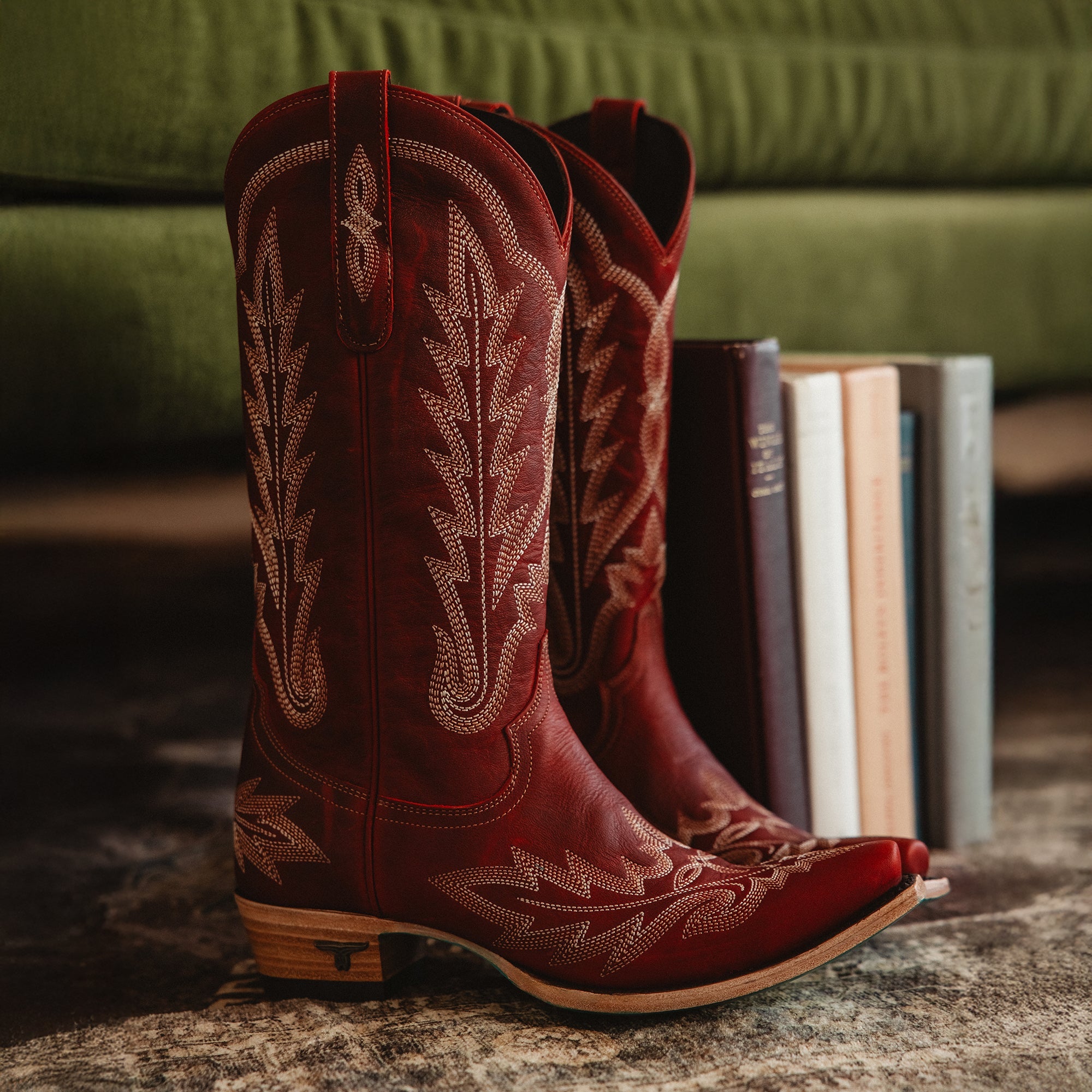 Pair of tall red cowboy boots with decorative stitching and wooden heels, placed on a rug beside a stack of books with a green couch in the background