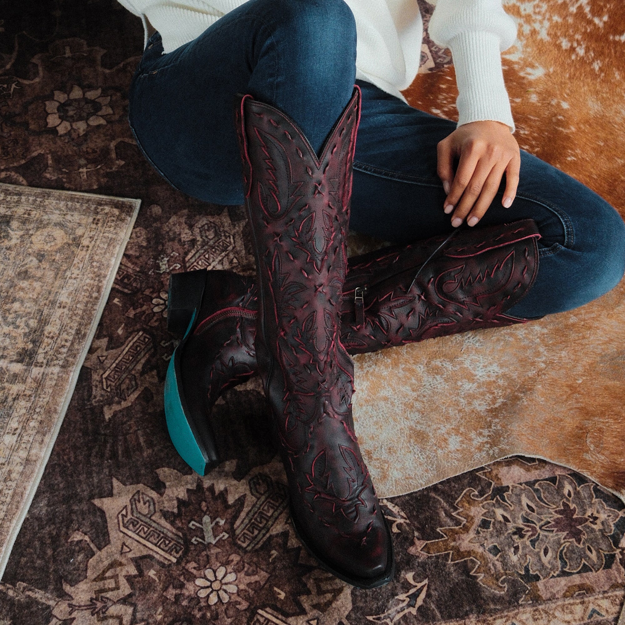 Woman sitting on a patterned rug wearing dark red cowboy boots with red stitching and turquoise soles, paired with dark jeans and a white sweater