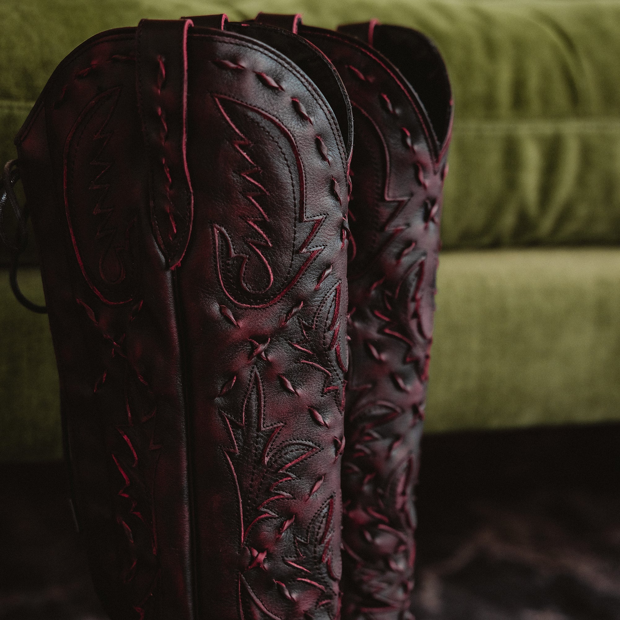 Close-up of the upper shafts of dark red cowboy boots with red stitching, positioned in front of a green couch