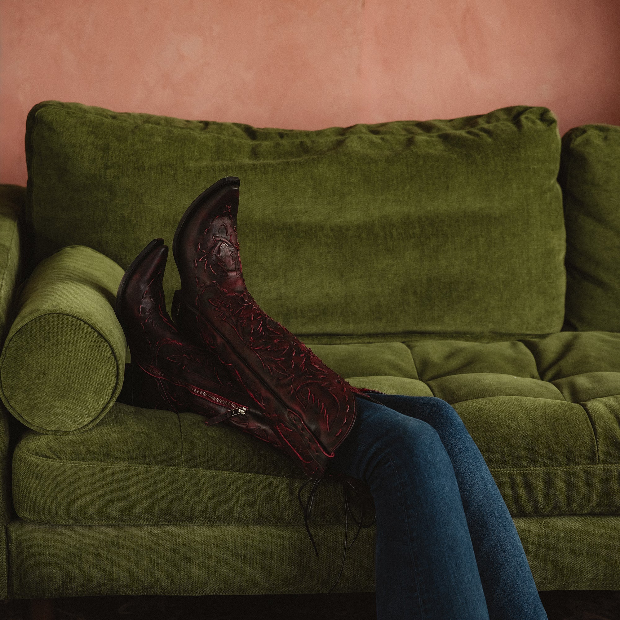 Person lying in front of a green couch with legs raised, wearing dark red cowboy boots with red stitching and dark jeans