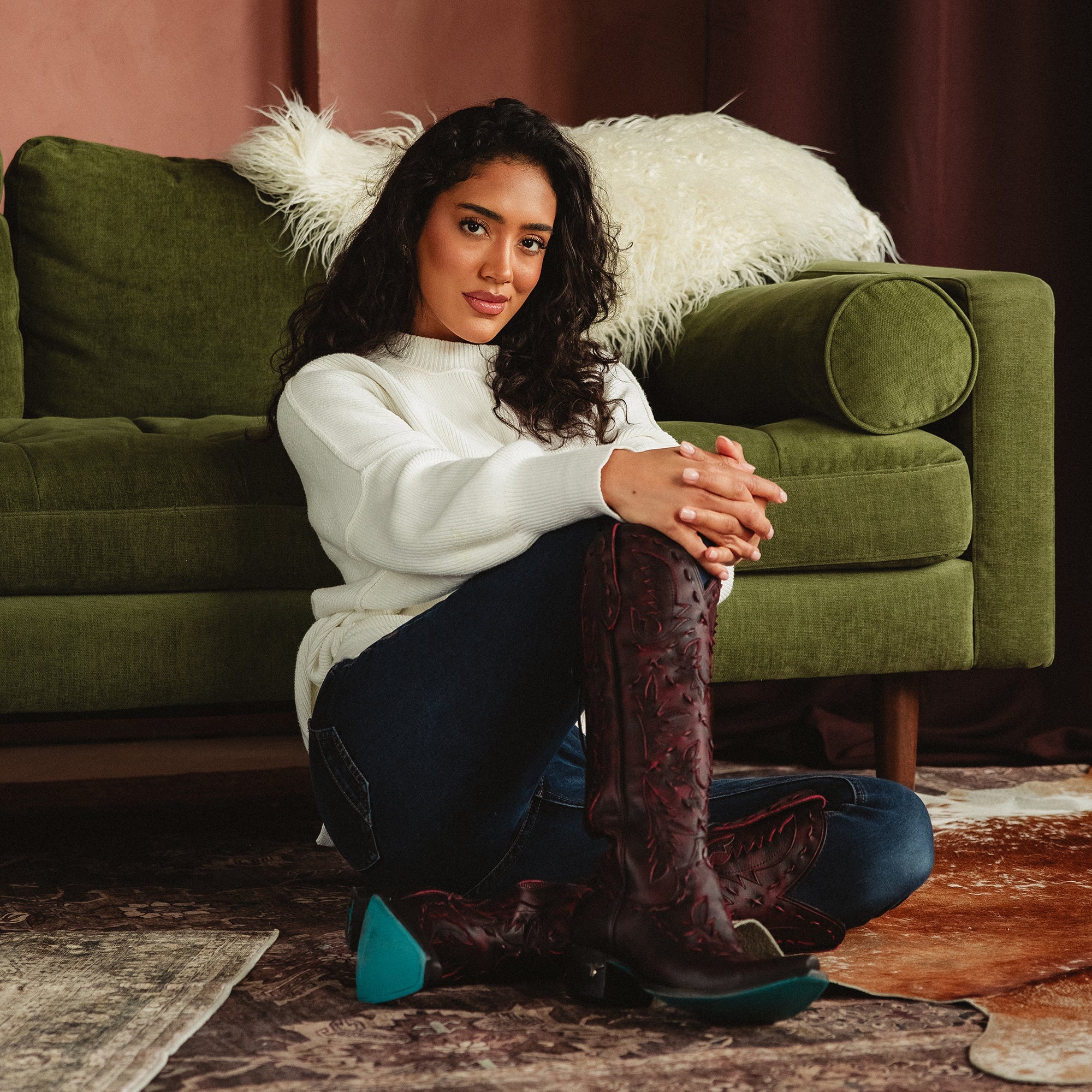 Woman sitting on the floor in front of a green couch wearing dark red cowboy boots with red stitching and turquoise soles, paired with dark jeans and a white sweater