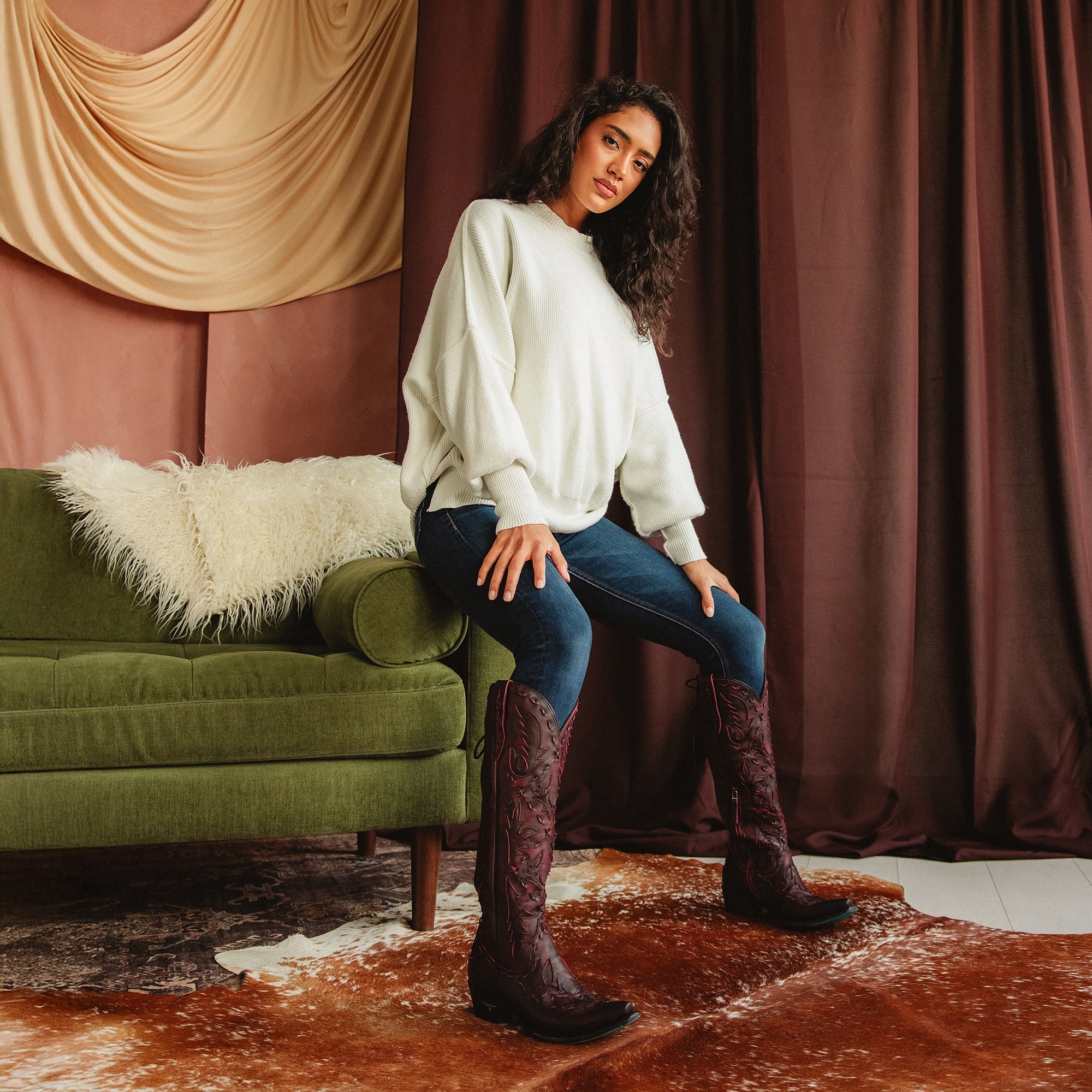 Woman sitting on the arm rest of a green couch wearing dark red cowboy boots with red stitching, paired with dark jeans and a white sweater