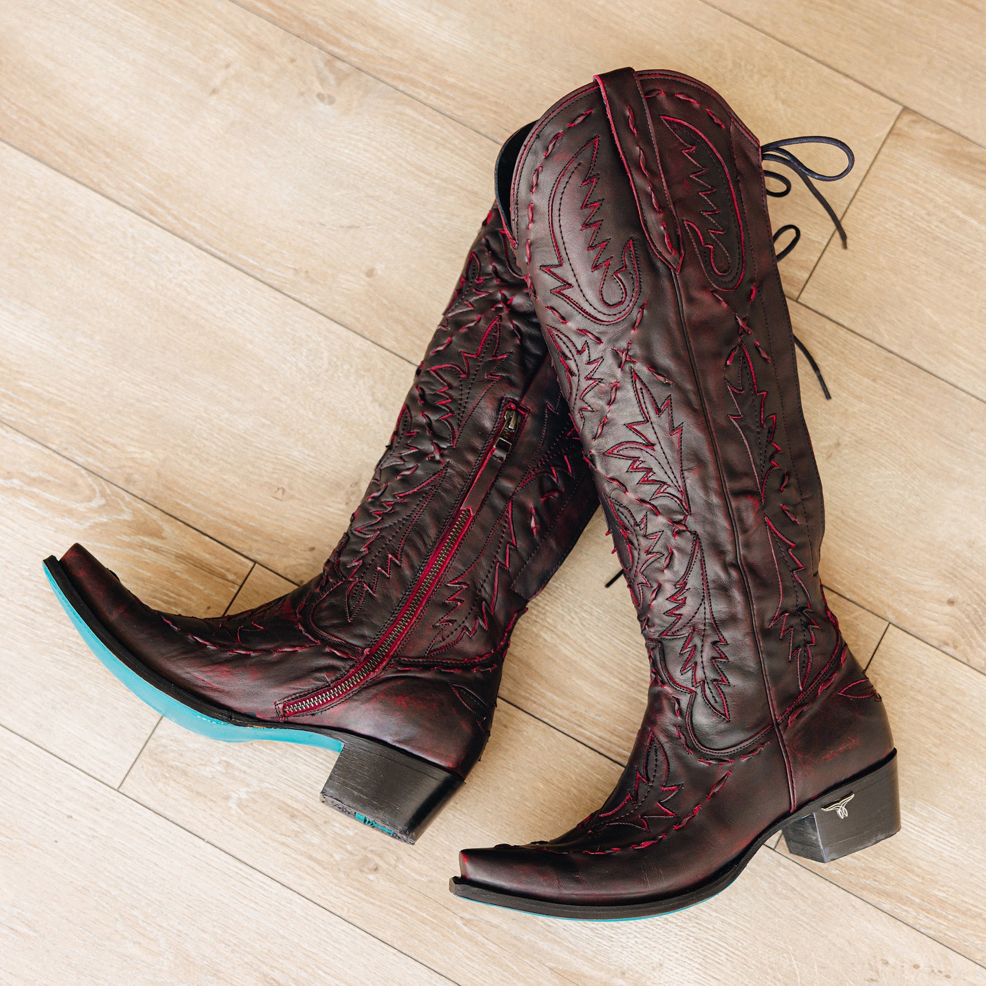 Pair of dark red cowboy boots with red stitching, black heels, turquoise soles, and side zippers, placed on a light wood floor
