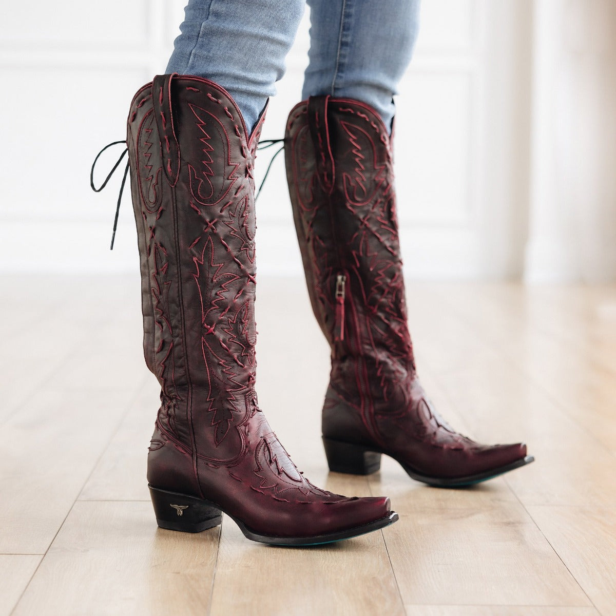 Person wearing dark red cowboy boots with red stitching and black heels, paired with light blue jeans on a wood floor