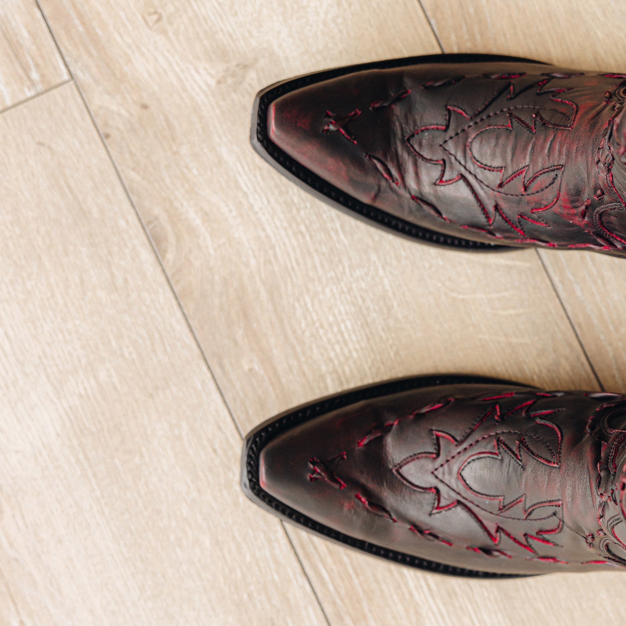 Close-up top view of dark red cowboy boots with red stitching and snip toes on a light wood floor