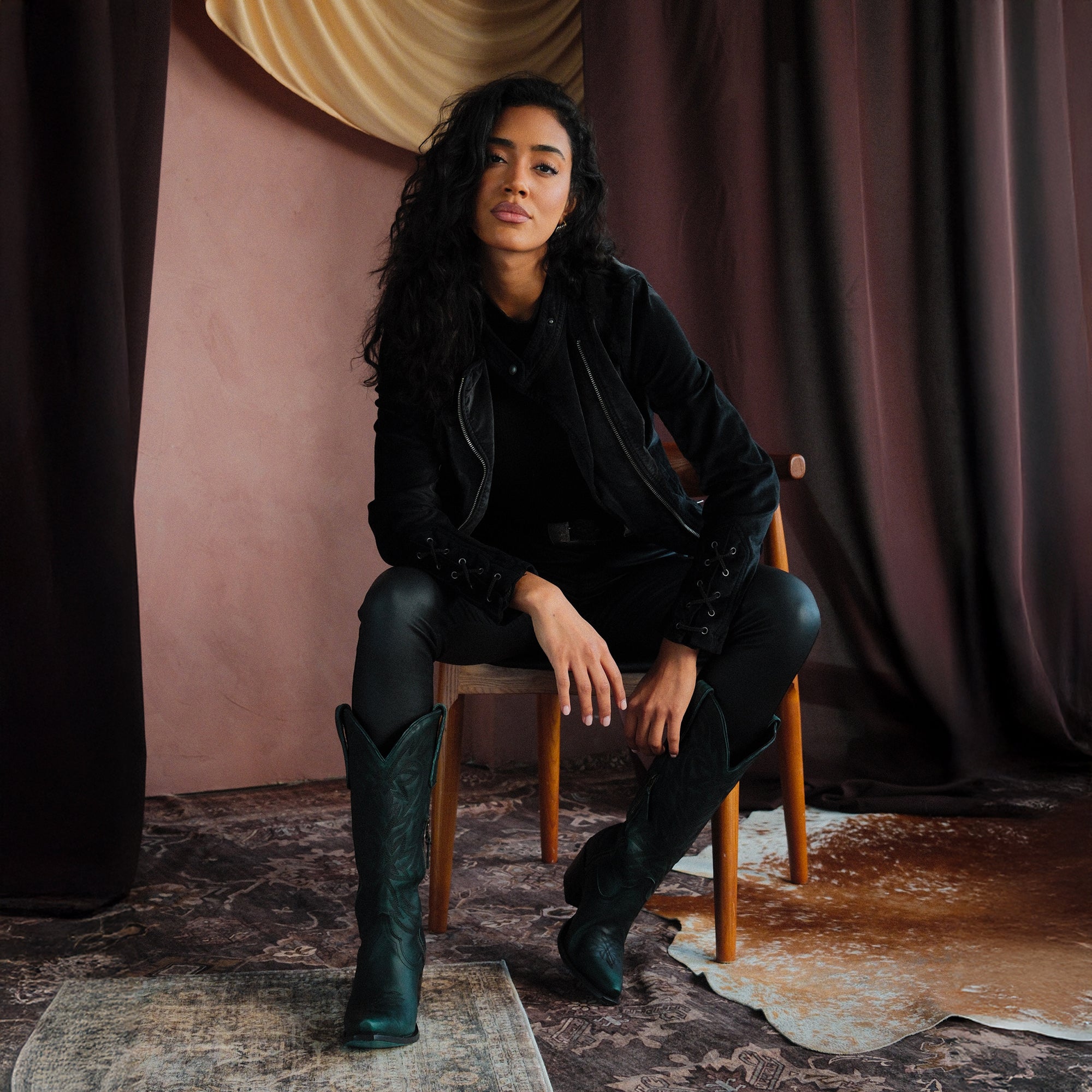 Woman sitting on a wooden chair wearing dark green cowboy boots, black pants, and a black jacket, posed against a dramatic draped backdrop