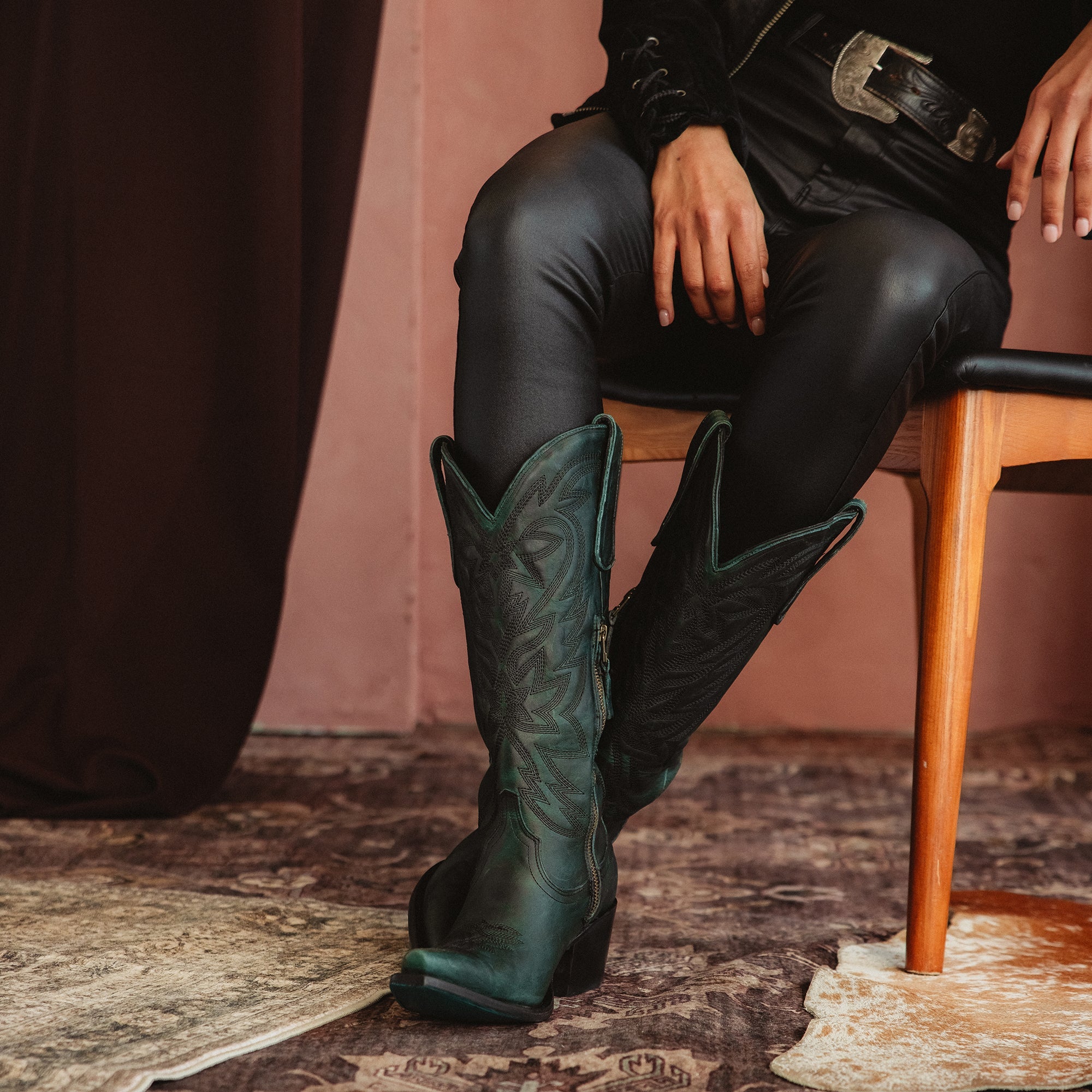 Woman sitting on a wooden chair wearing dark green cowboy boots, black pants, and a black jacket, posed against a dramatic draped backdrop