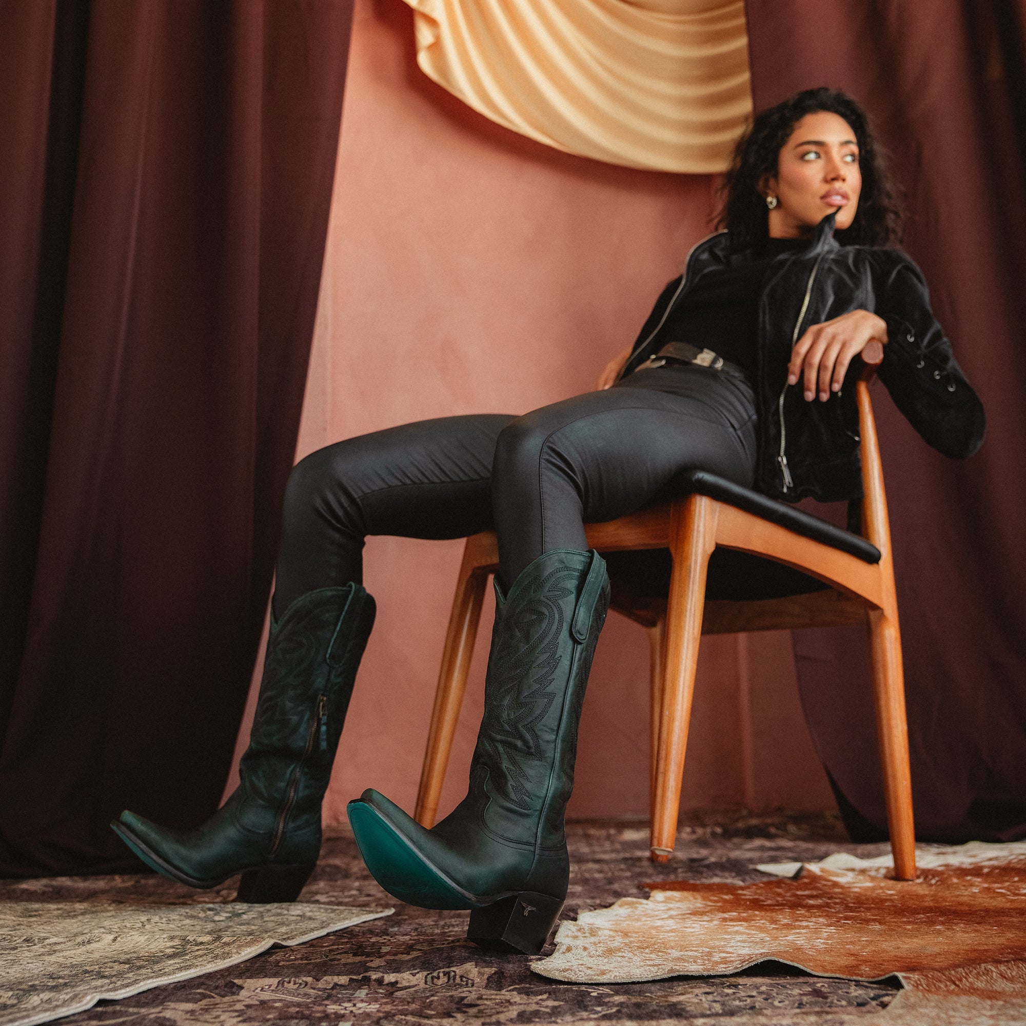 Woman sitting on a wooden chair wearing dark green cowboy boots with turquoise soles, black pants, and a black jacket, posed against a dramatic draped backdrop