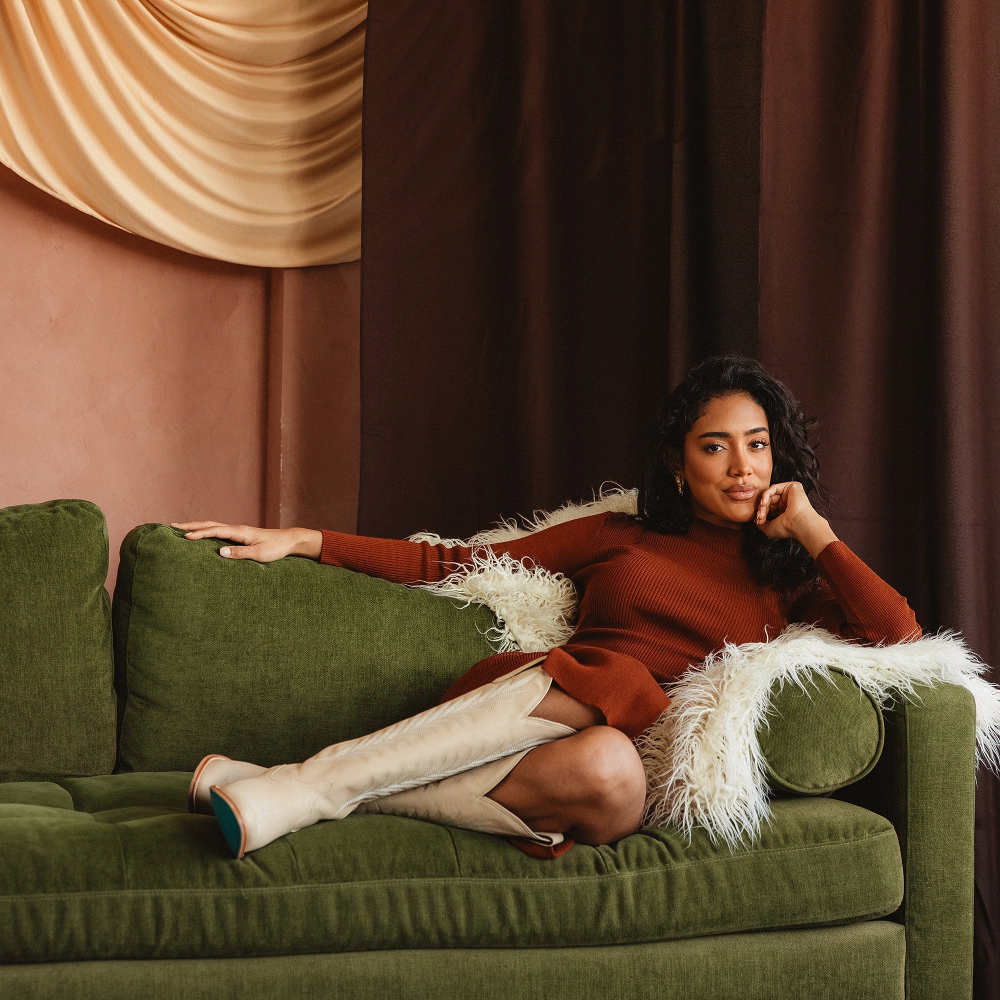 Woman lounging on a green couch wearing tall cream-colored cowboy boots with a stitched design and pointed toes, paired with a rust-colored outfit