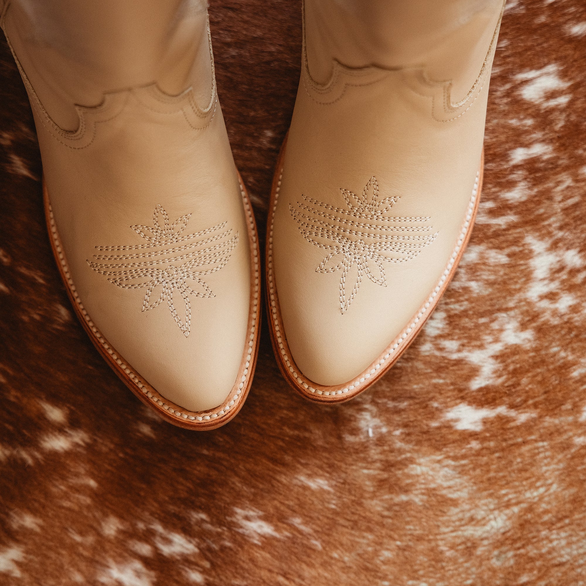 Close-up of cream-colored cowboy boots with decorative stitching on the toes, resting on a brown and white cowhide rug