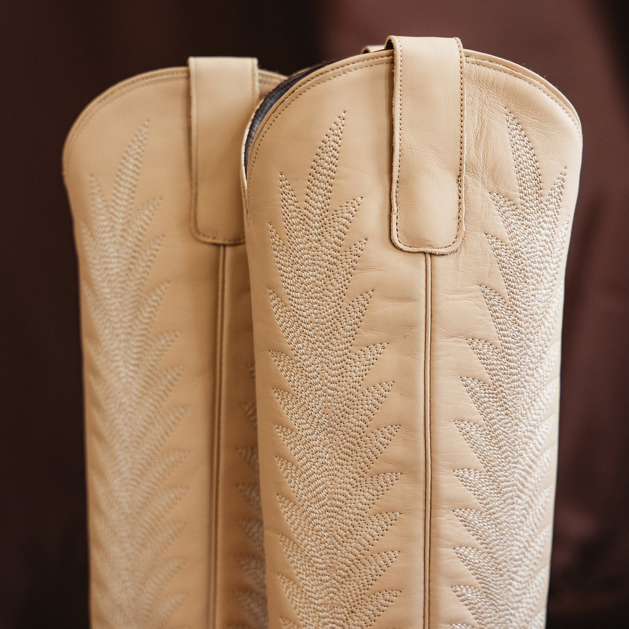 Close-up of the top portion of cream-colored cowboy boots featuring detailed stitched patterns and sturdy pull straps