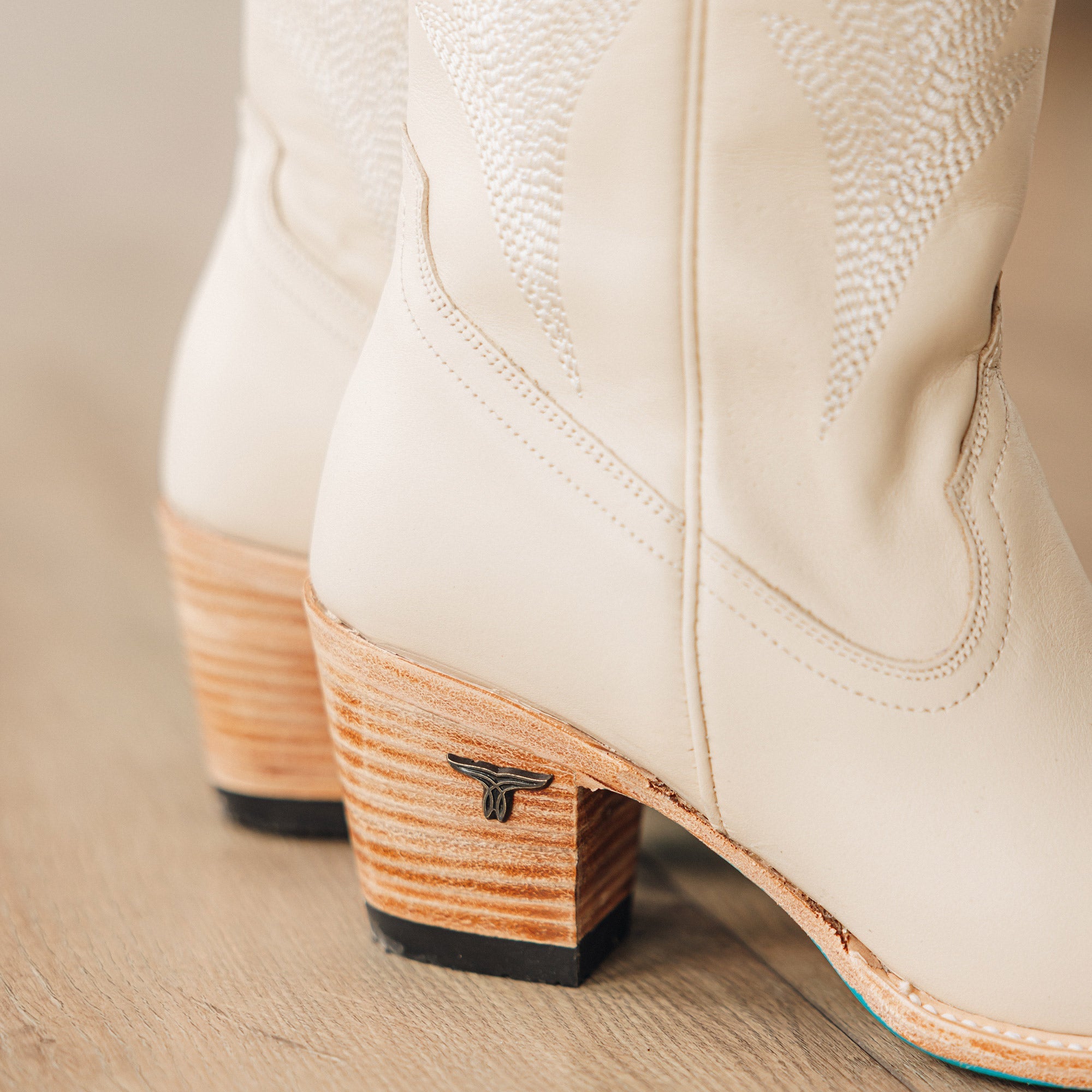 Close-up of cream-colored cowboy boots focusing on the stacked wooden heels with a small bull head emblem and curved stitching along the sides