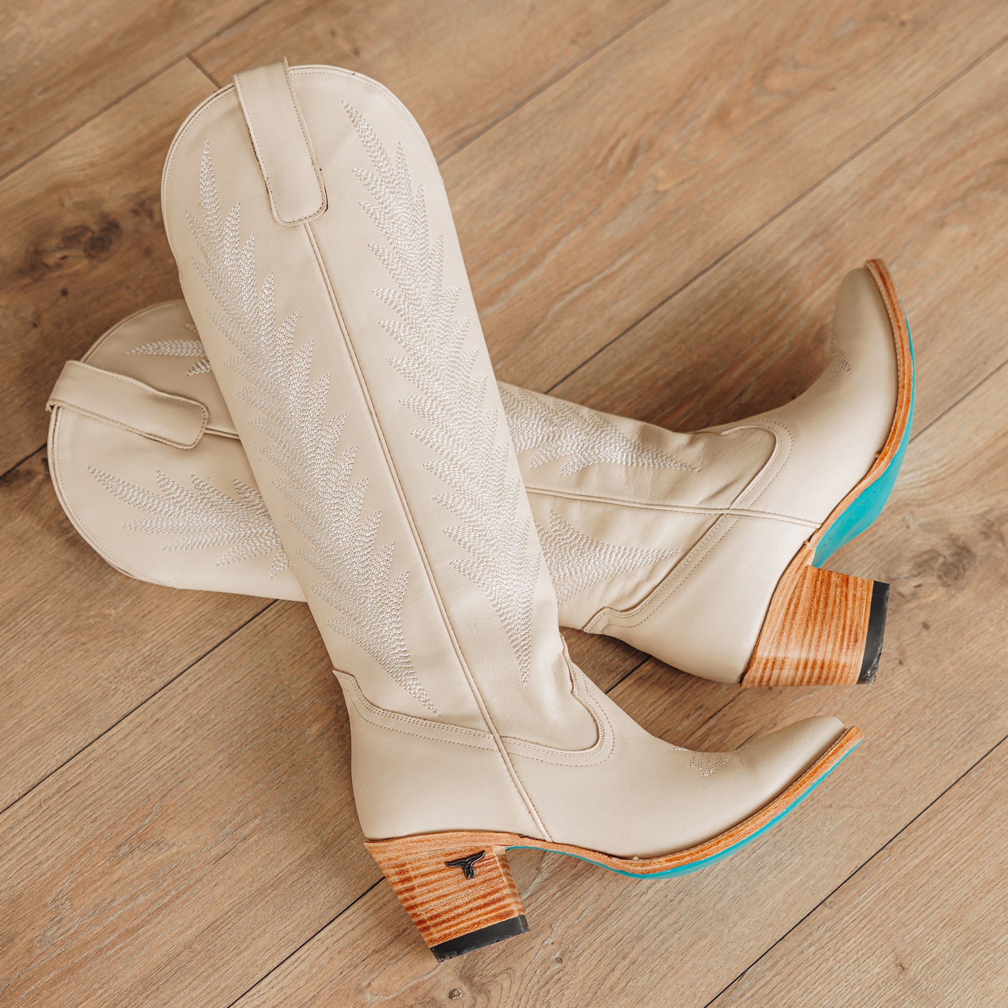 Pair of tall cream colored cowboy boots with stitched leaf patterns along the shaft, wooden stacked heels, and turquoise soles, displayed on a light wood floor