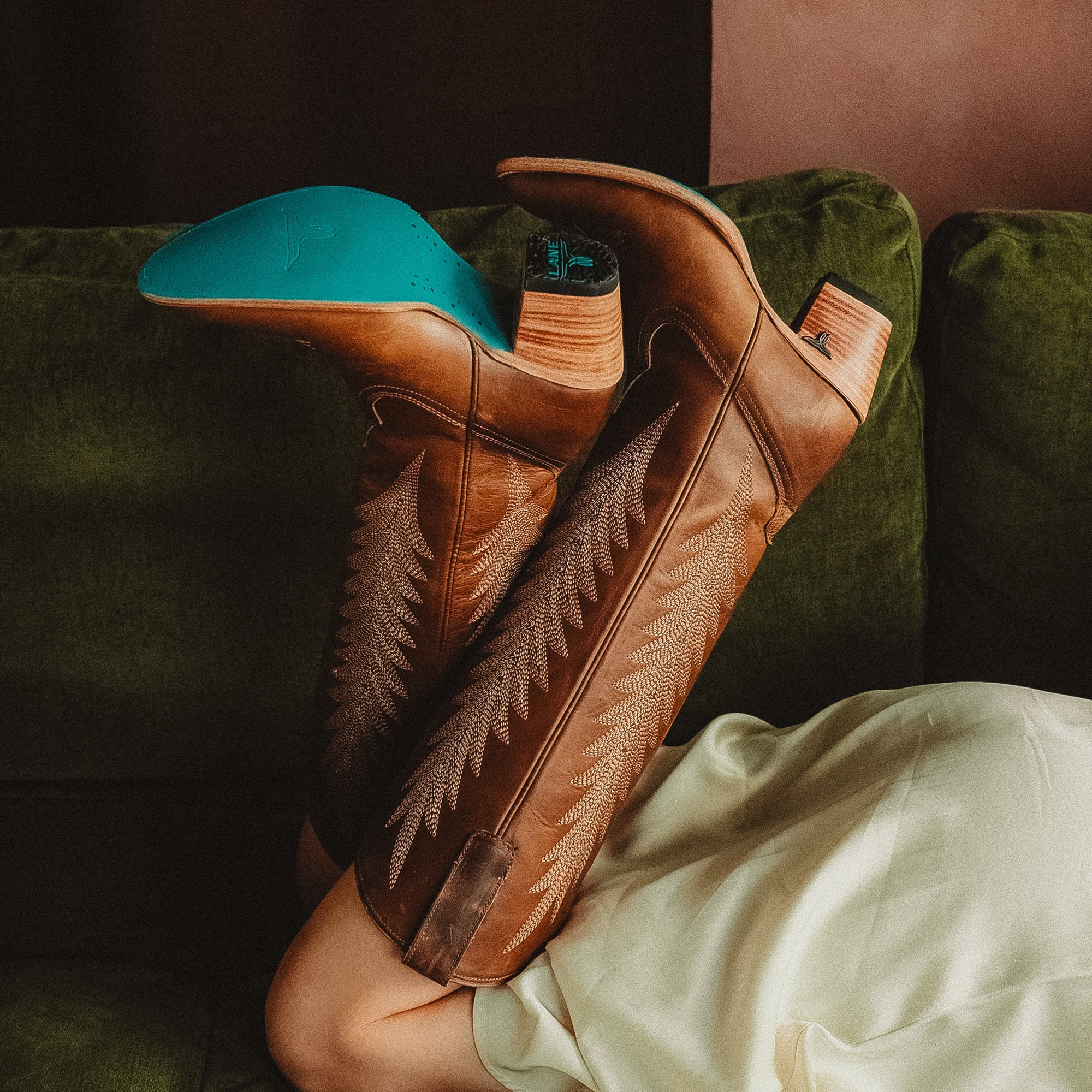 Woman lying on a green couch with legs raised, wearing tall brown cowboy boots featuring decorative stitching and turquoise soles