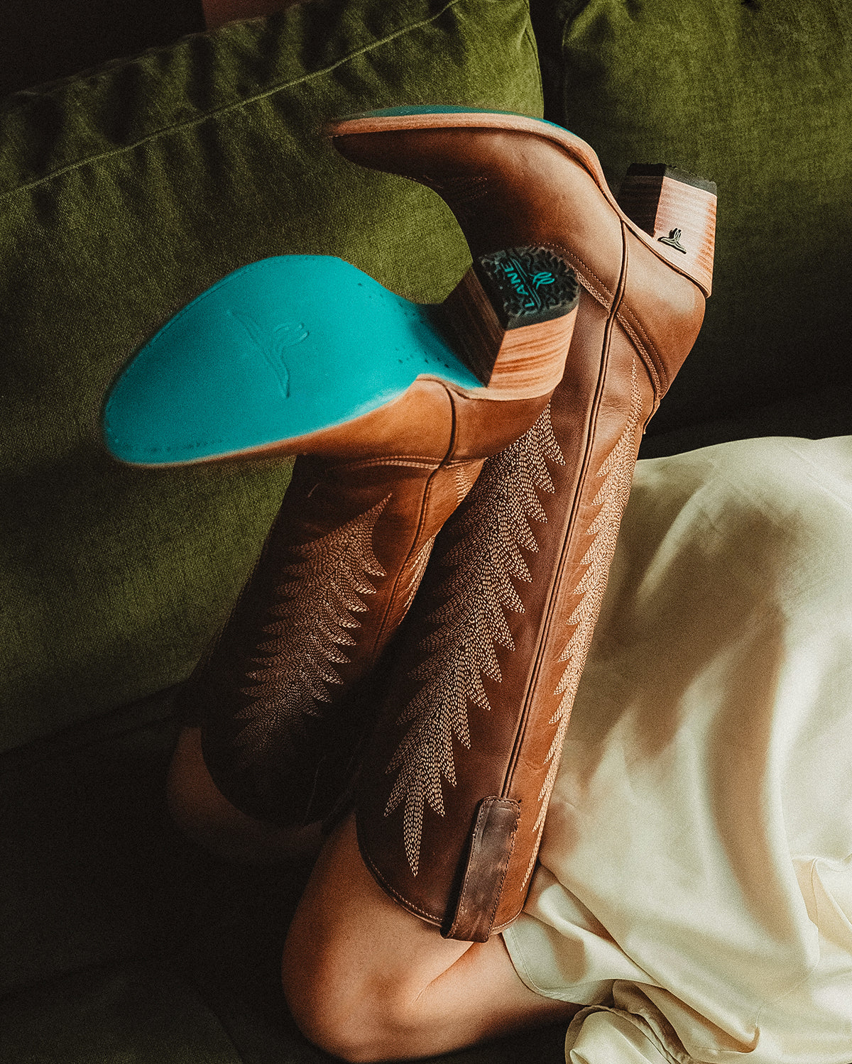 Person lying on a couch with legs raised, wearing tall brown cowboy boots with decorative stitching and turquoise soles