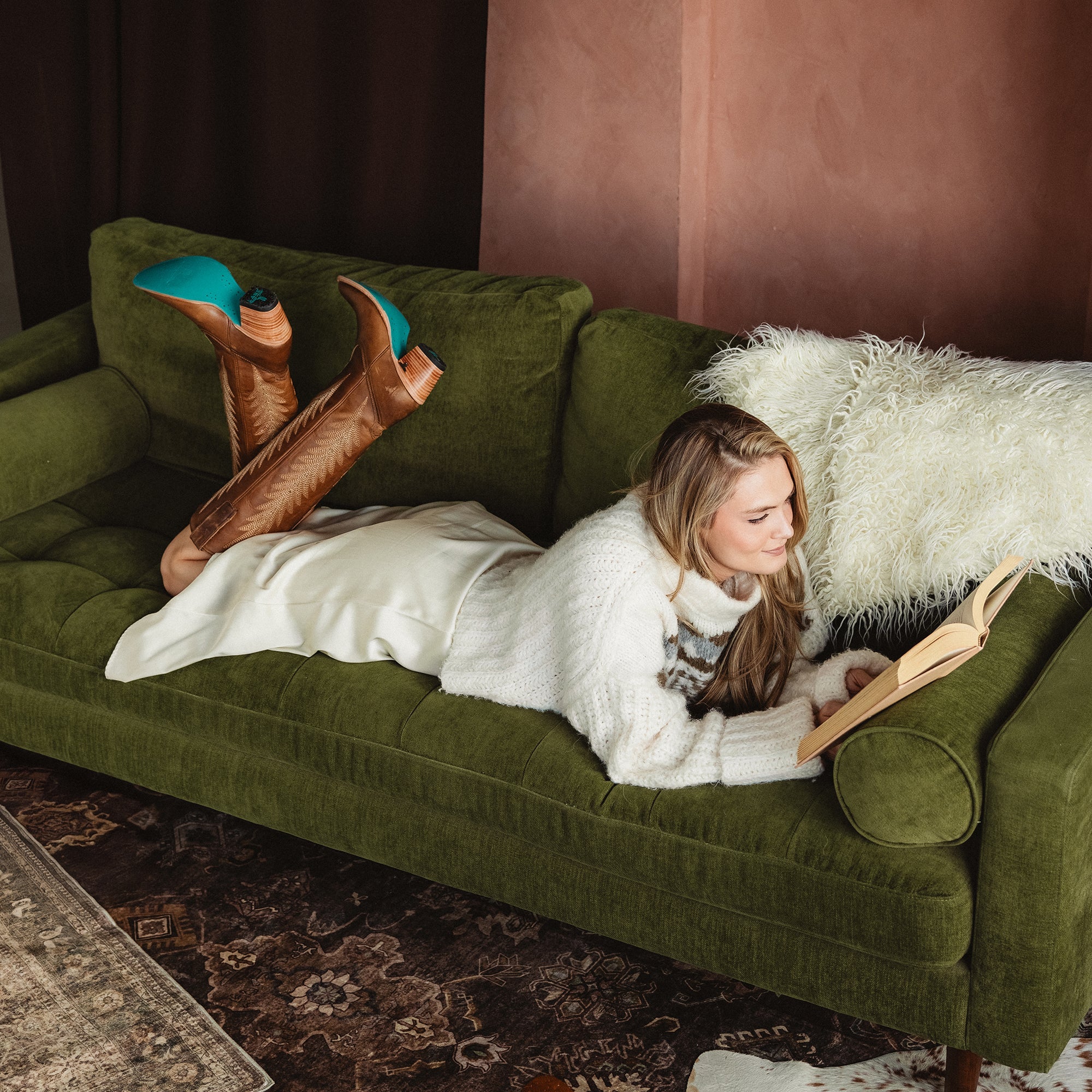 Woman lying on a green couch reading a book, wearing a white sweater and skirt with tall brown cowboy boots that have turquoise soles, with her legs playfully raised behind her