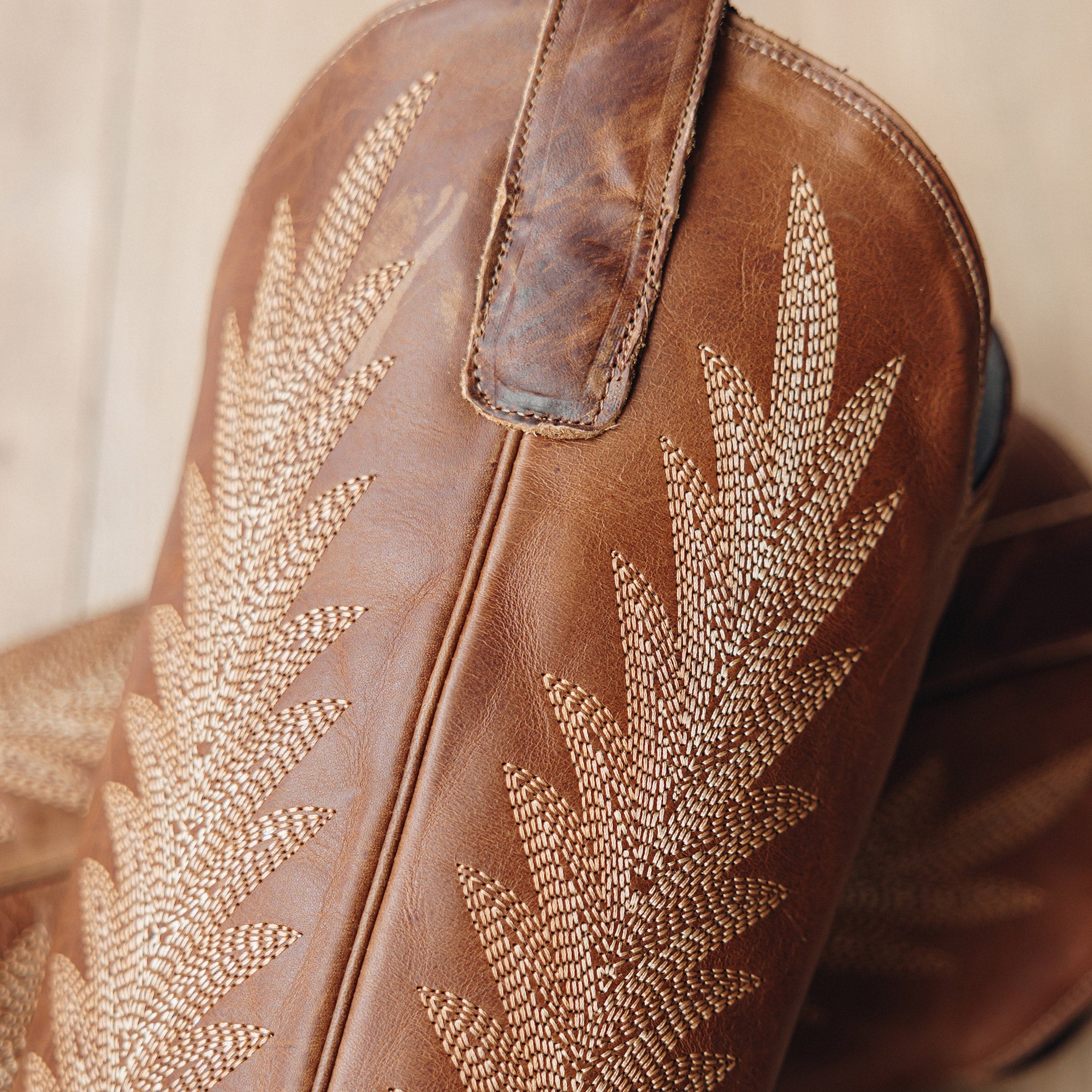 Close-up of brown leather cowboy boots featuring intricate stitched leaf patterns along the shaft and a sturdy pull strap at the top