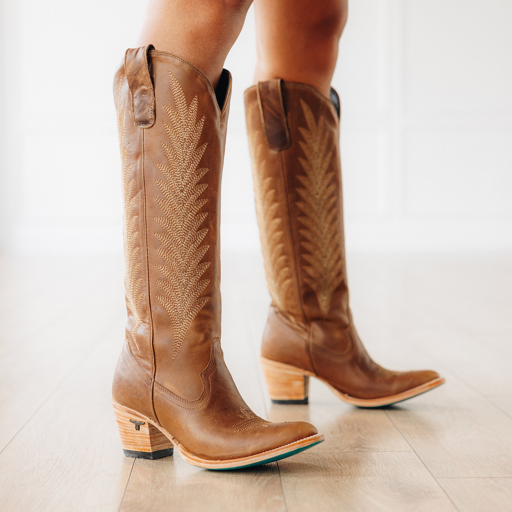 Close-up side view of a person wearing tall brown cowboy boots with decorative stitching, wooden stacked heels, and turquoise soles, standing on a light wood floor