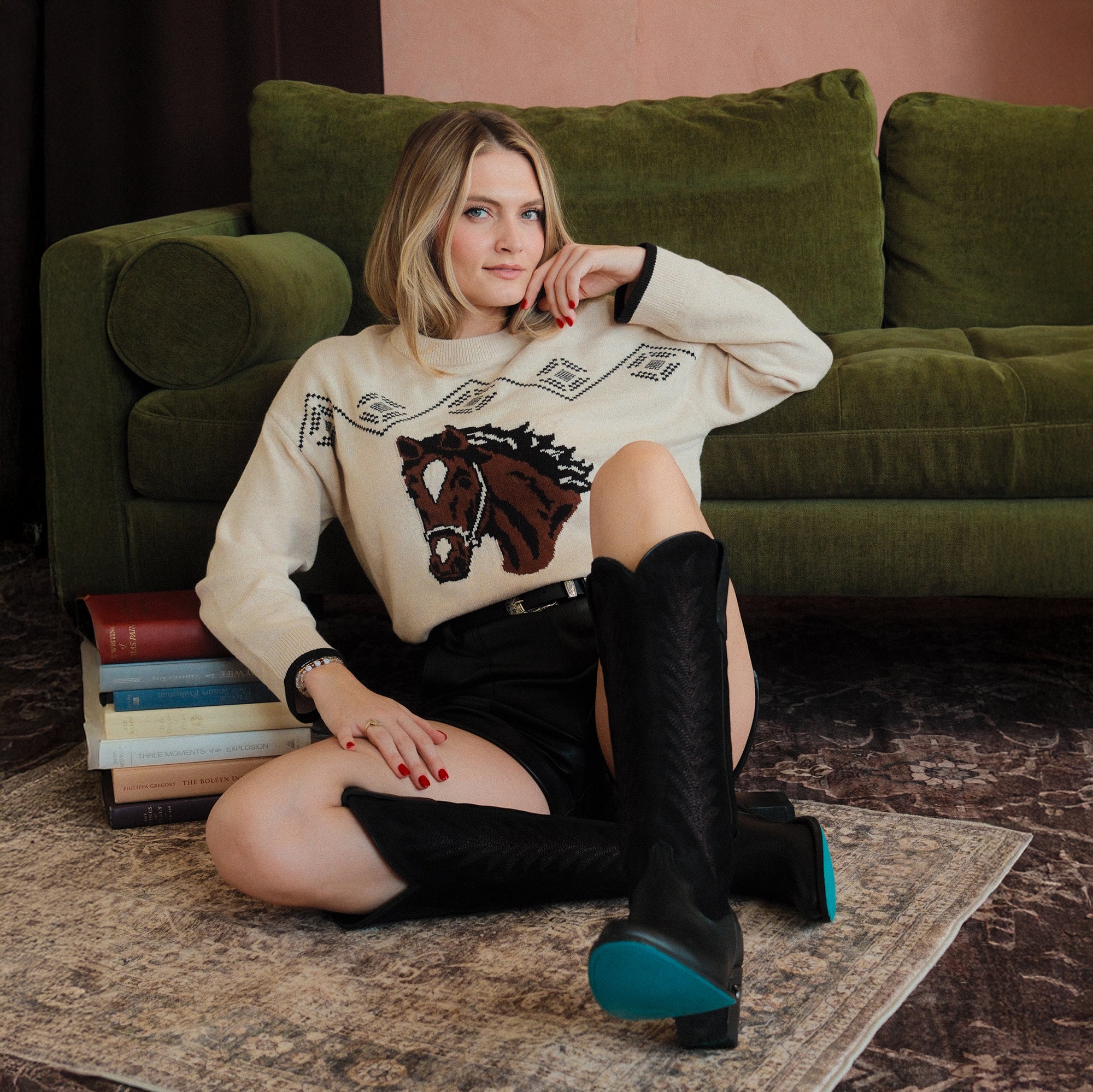 Woman sitting on the floor in front of a green couch, wearing black cowboy boots with turquoise soles and a cream sweater featuring a horse graphic. A stack of books is positioned to the side