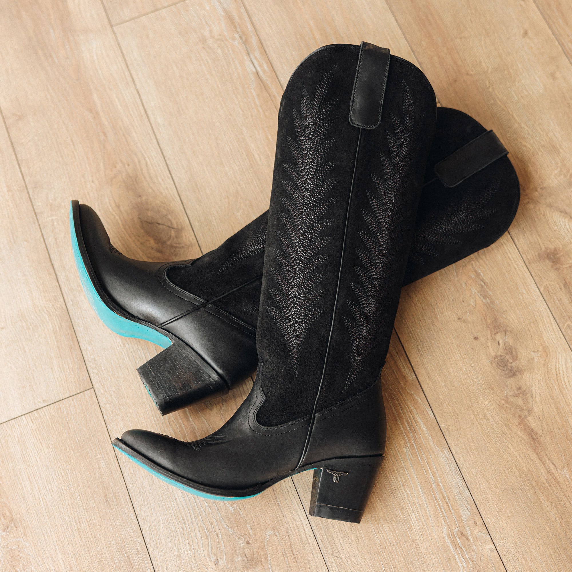 Pair of black cowboy boots with stitched detailing and turquoise soles, displayed on a light wood floor with one boot resting over the other