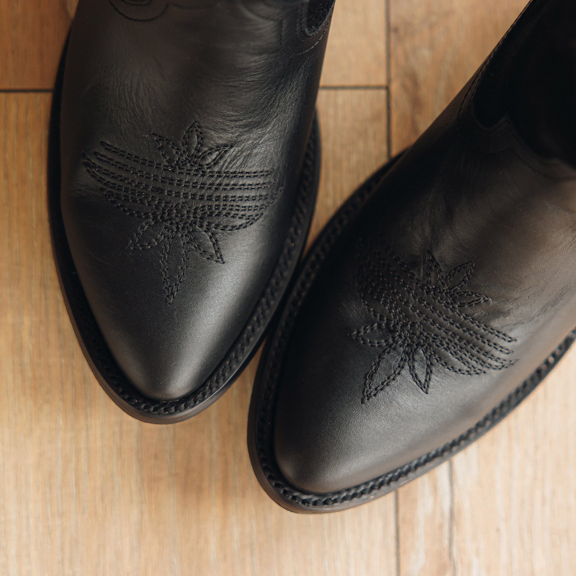 Overhead view of a pair of black cowboy boots on a wood floor, showing decorative stitched patterns on the toes