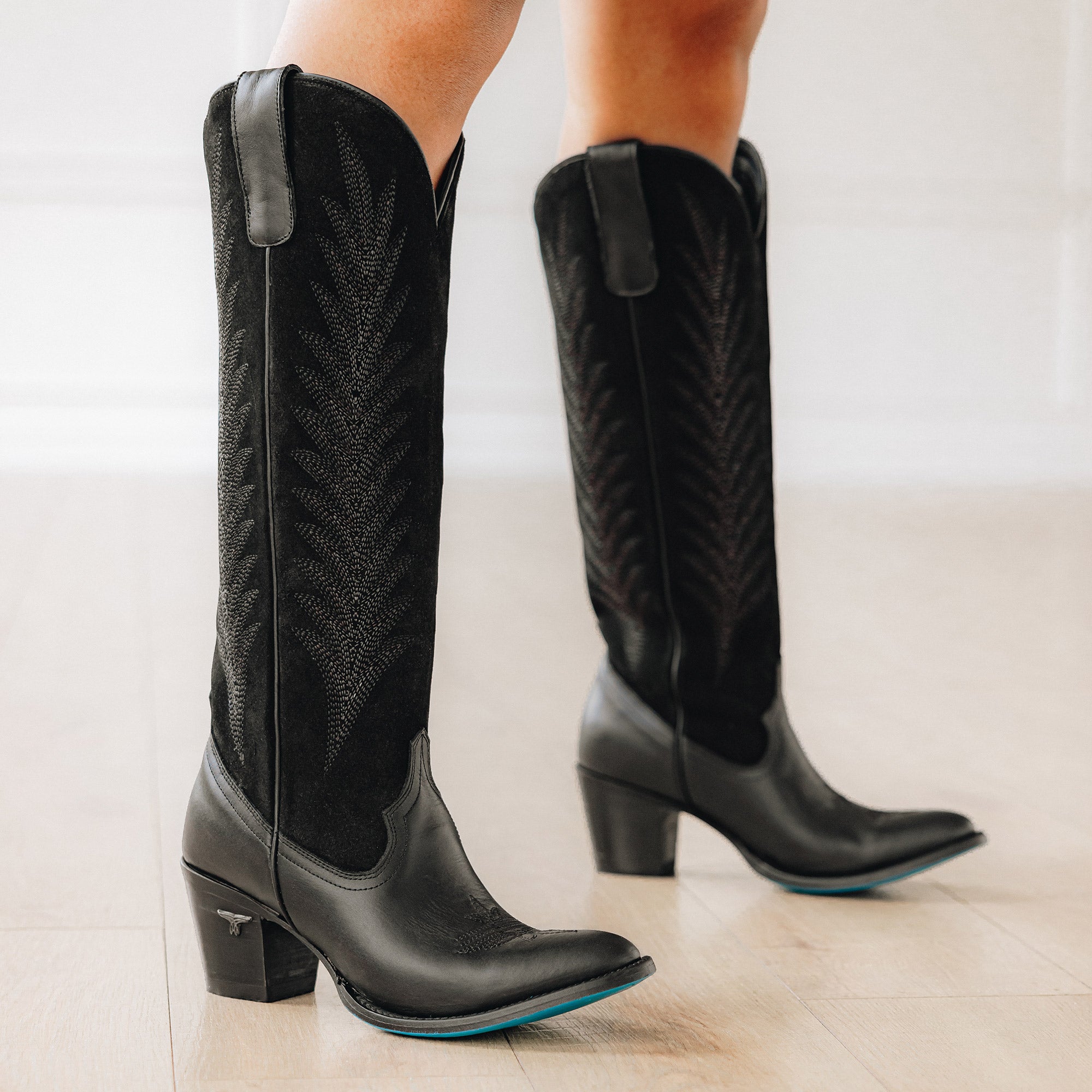 Close-up of a person standing indoors wearing tall black cowboy boots with stitched detailing and turquoise soles on light wood flooring