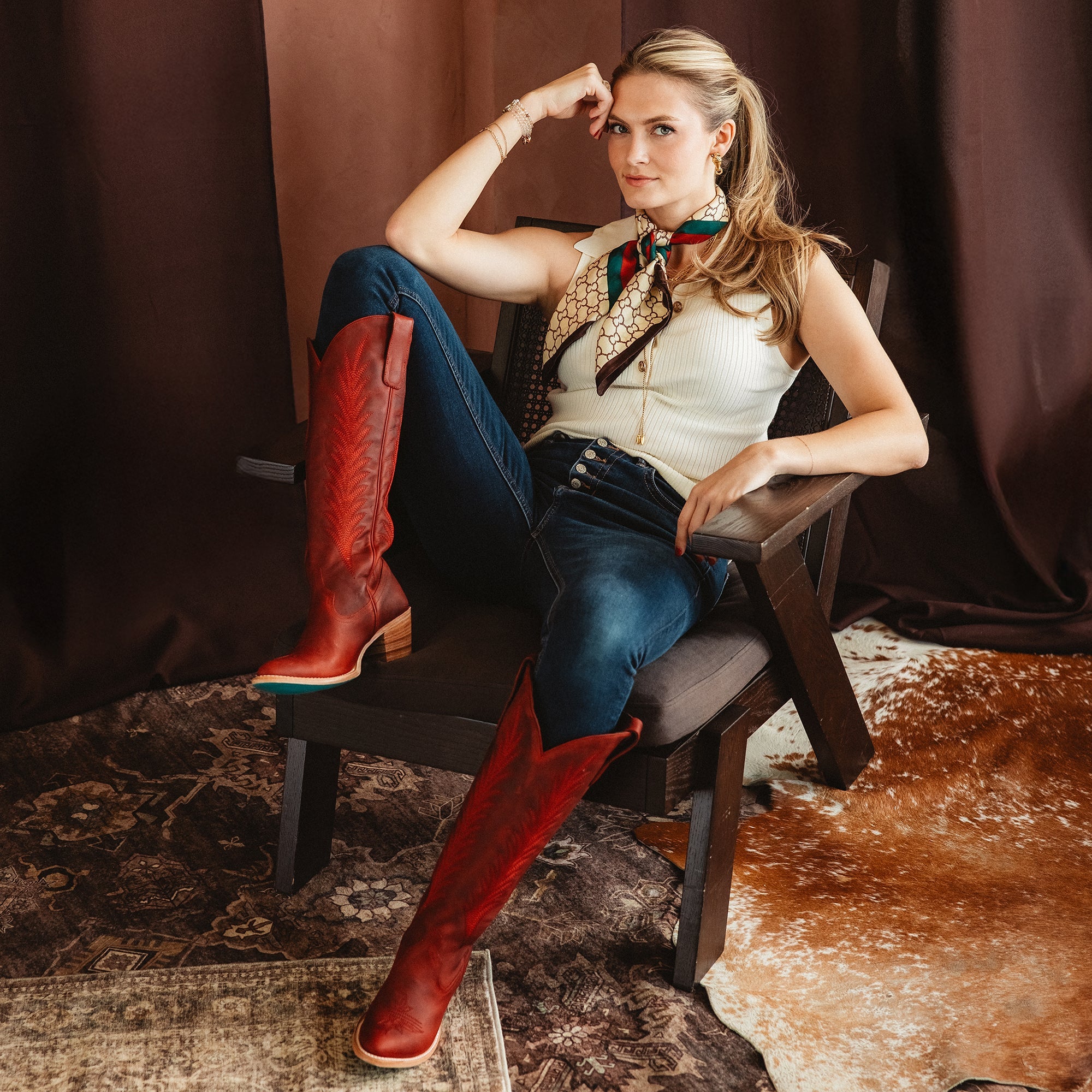 Woman sitting in a chair wearing tall red cowboy boots with decorative stitching, dark jeans, a sleeveless top, and a patterned scarf, posed on a patterned rug with a dark curtain backdrop