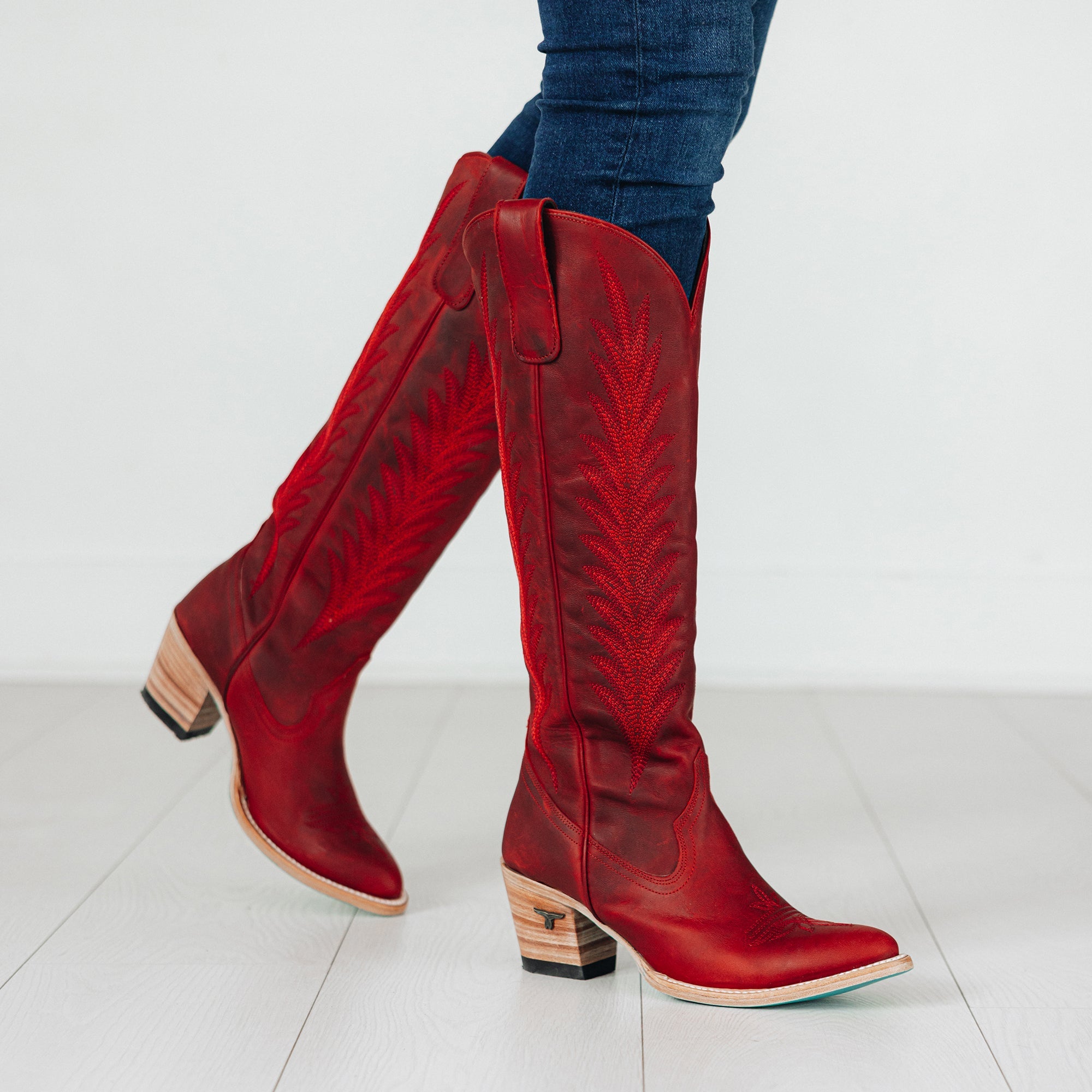 Tall red cowboy boots with decorative stitching and stacked wooden heels, worn with dark jeans on a white floor background