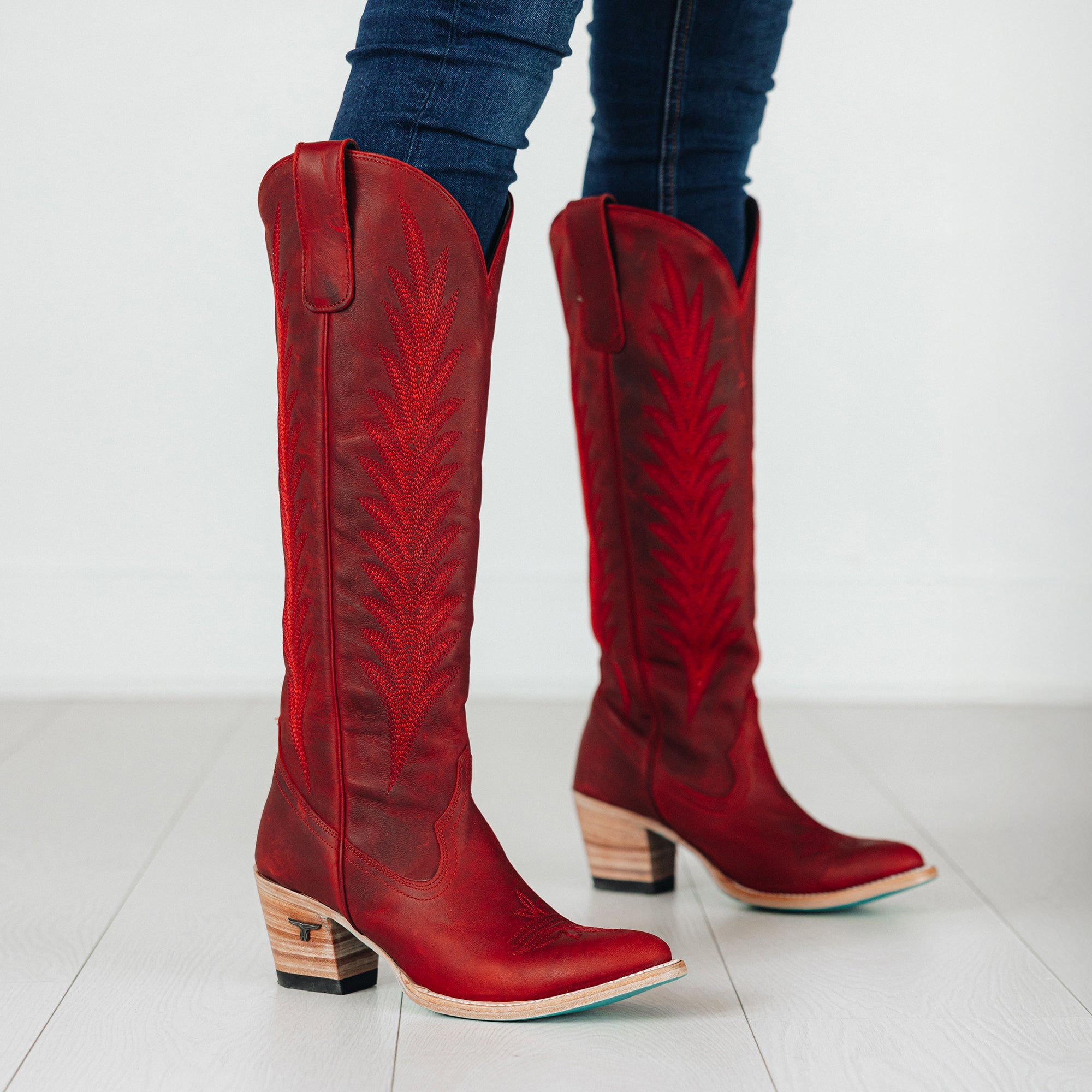 Tall red cowboy boots with decorative stitching and stacked wooden heels, worn with dark jeans on a white floor background